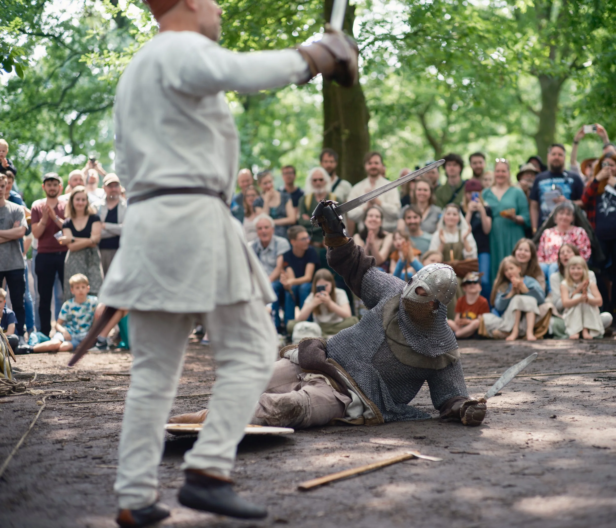A reenactment of a medieval or Renaissance combat scene in a wooded outdoor setting, with two men dressed in period costumes engaged in sword fighting while a crowd of spectators, including children, watches and records the event.