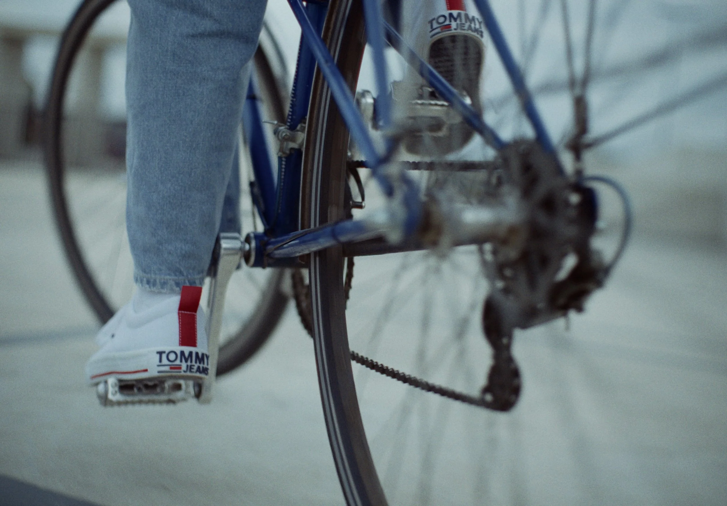 Close-up of a person's leg in light blue jeans and white sneakers with red and blue logo, riding a bicycle with a blue frame and a Tommy Hilfiger logo, on a road or indoor track.