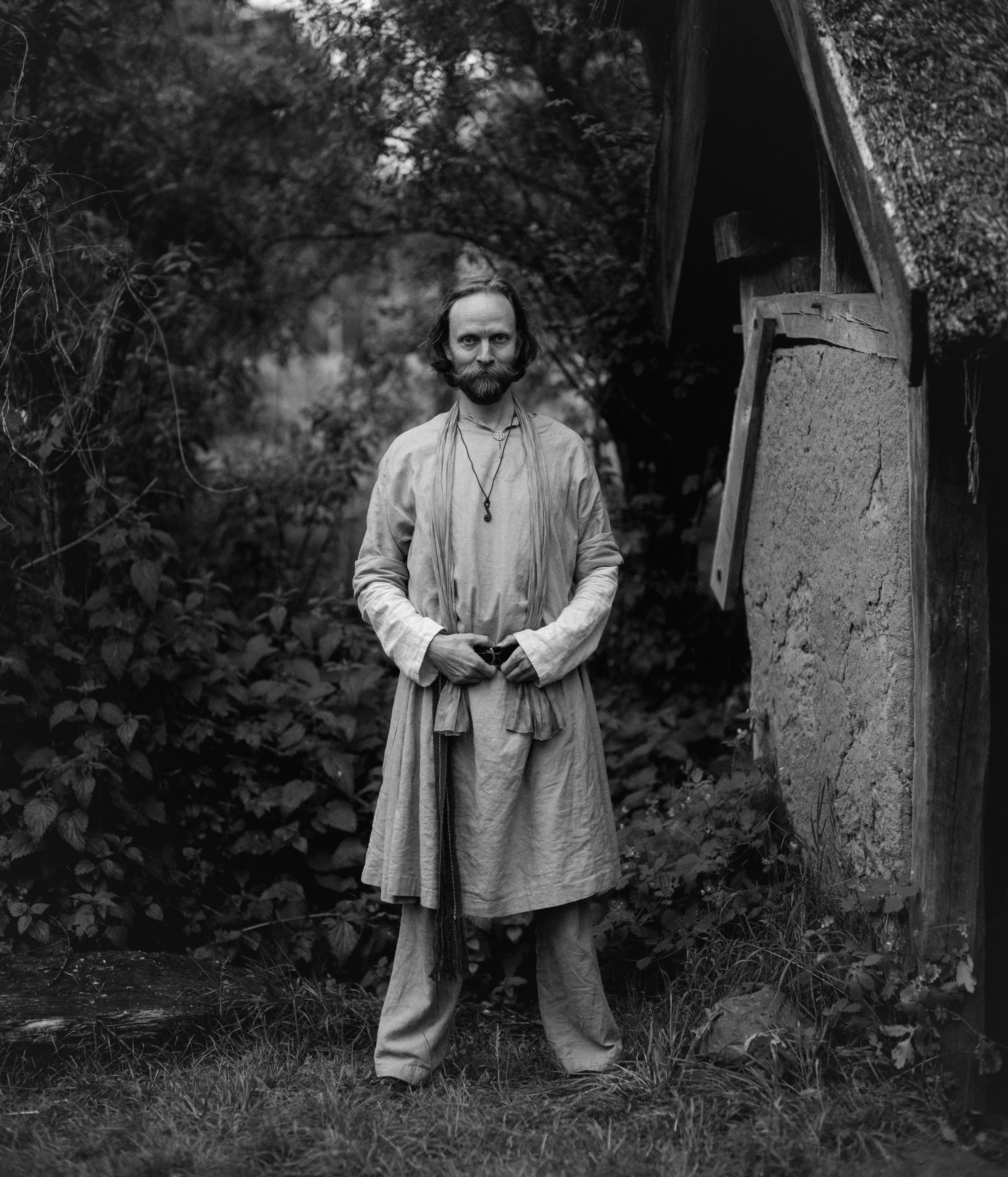 A man with a beard and long hair stands outdoors surrounded by trees and plants. He is wearing loose, light-colored clothing with a necklace, and holds something in his hands. There is a wooden structure or shed behind him on his right side.