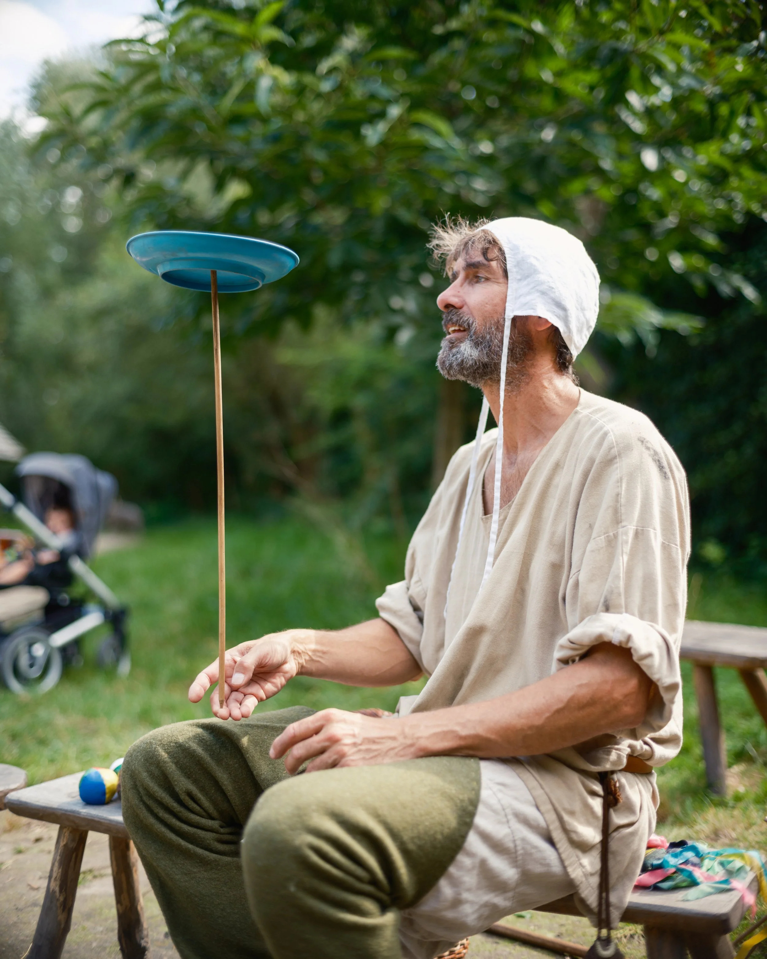 An older man with gray hair and beard sitting outdoors on a wooden bench, wearing a light-colored shirt and dark pants, balancing a blue spinning top on a stick in front of him, with trees and greenery in the background.