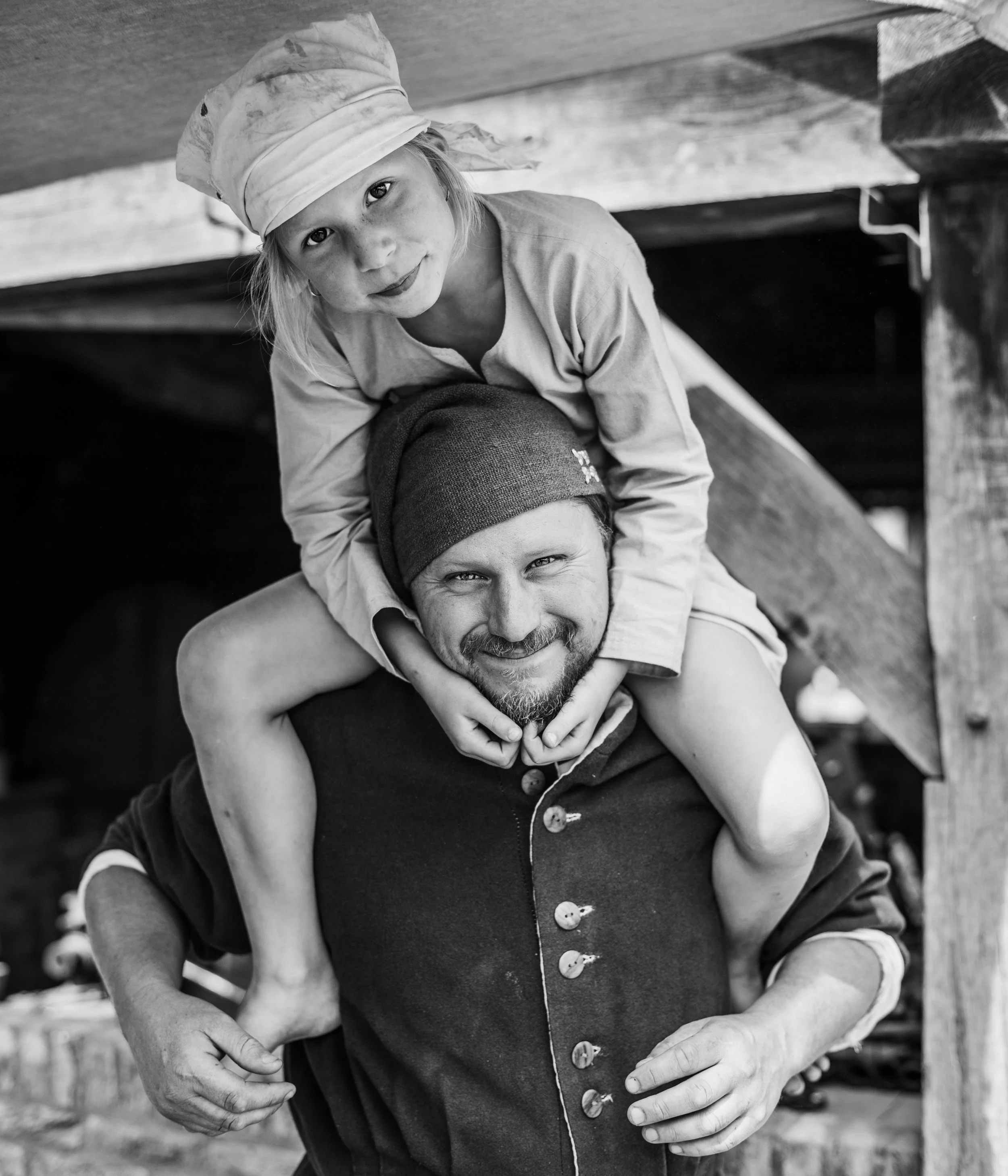 A black and white photo of a young girl wearing a chef's hat and a jacket with large buttons, leaning over and holding a man's shoulders. They are both smiling, and the setting appears to be outdoors under a wooden structure.