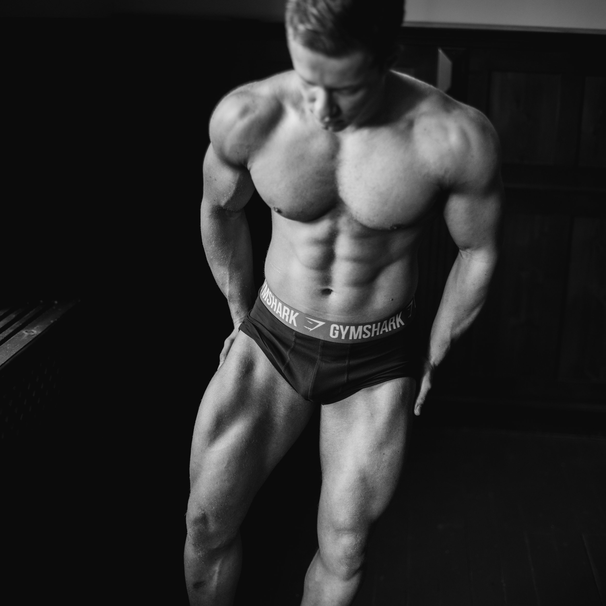 Black and white photo of a shirtless man with a muscular build, wearing gym shorts with the brand Gymshark, standing in a dark room with his head looking down.