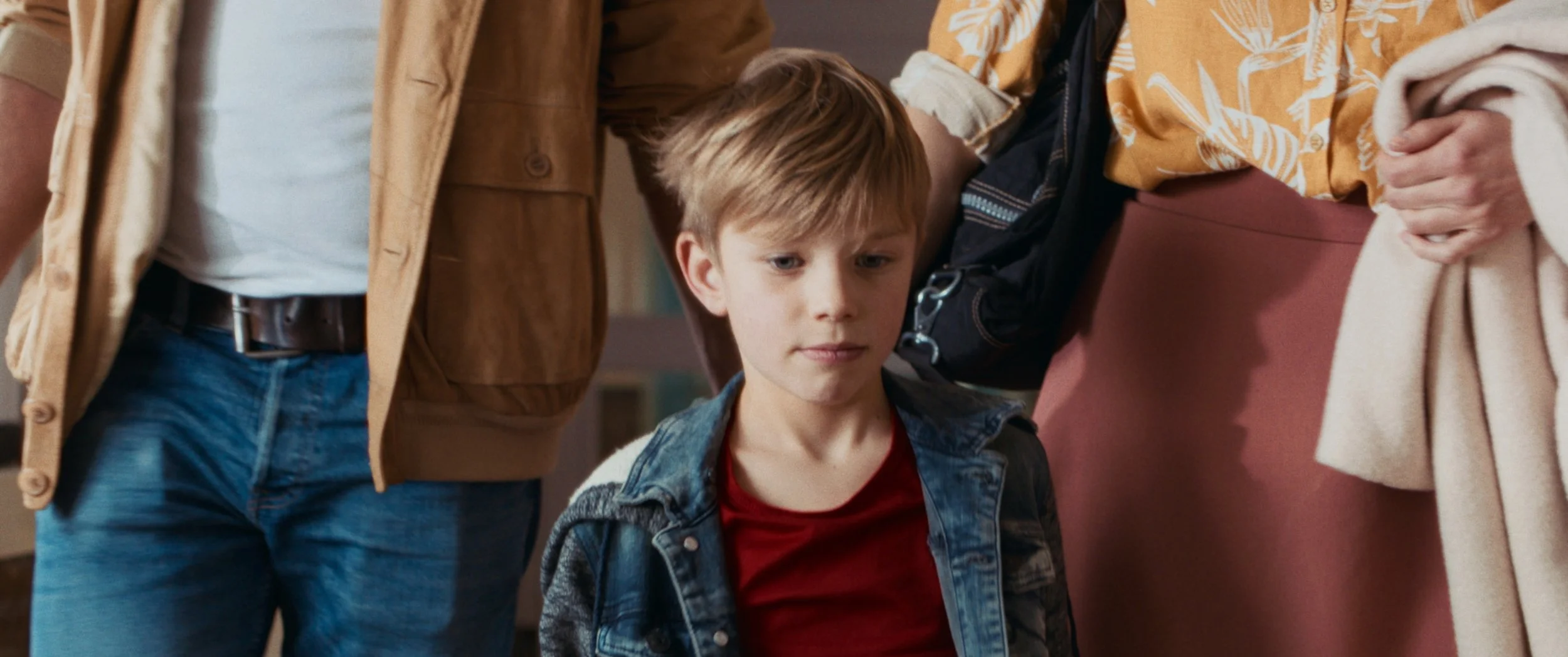 A young boy with light brown hair wearing a denim jacket and red shirt, looking down with a neutral expression, surrounded by adults.