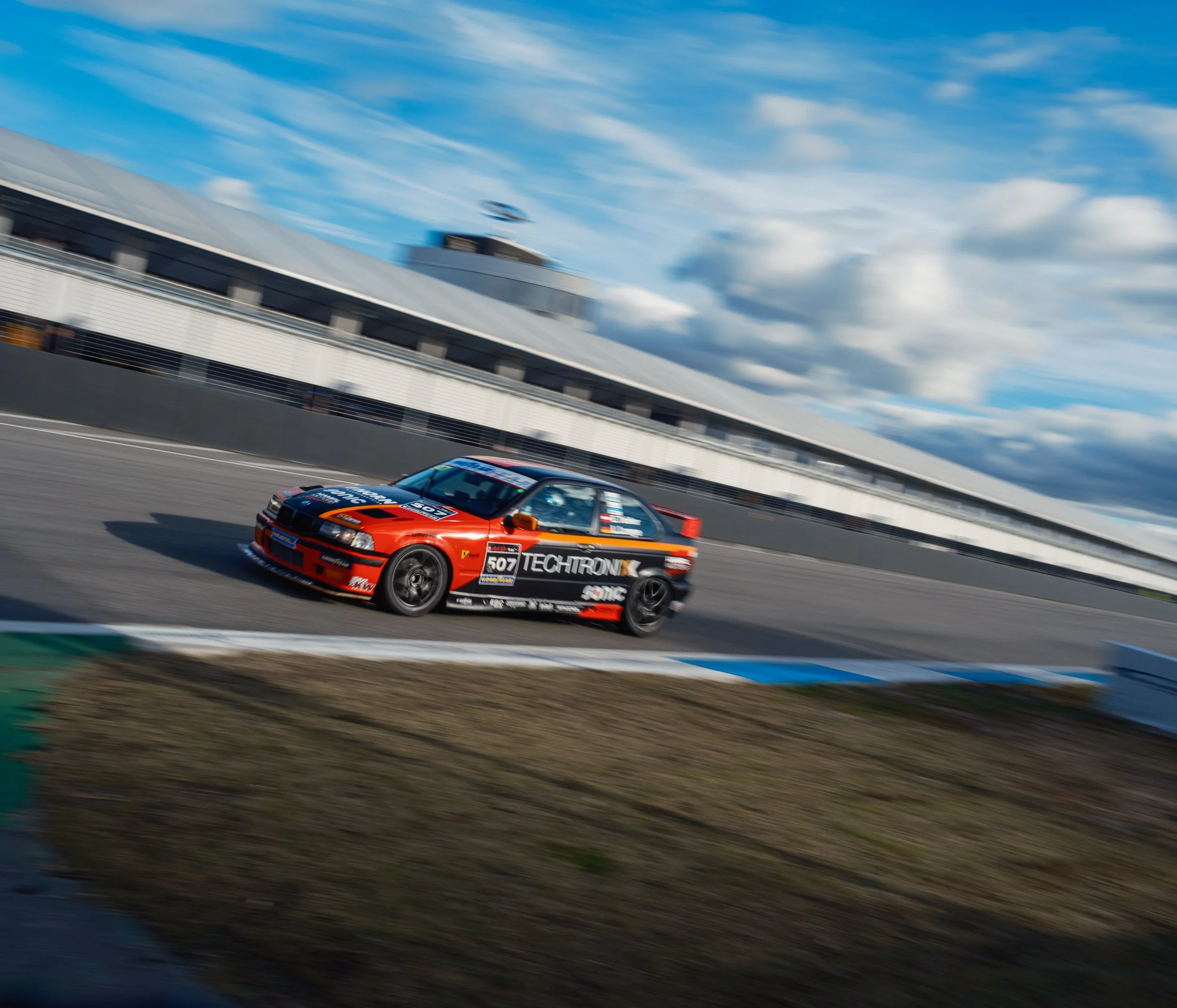 A race car on a race track during daytime, with a modern building and a blue sky with clouds in the background.