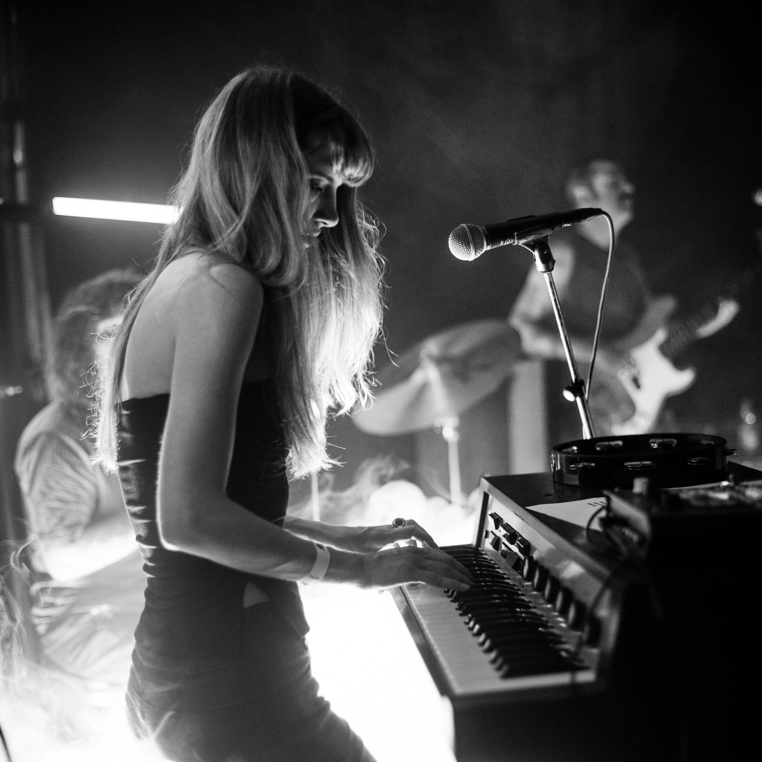 A woman with long hair playing a piano in a dark setting with bright background light
