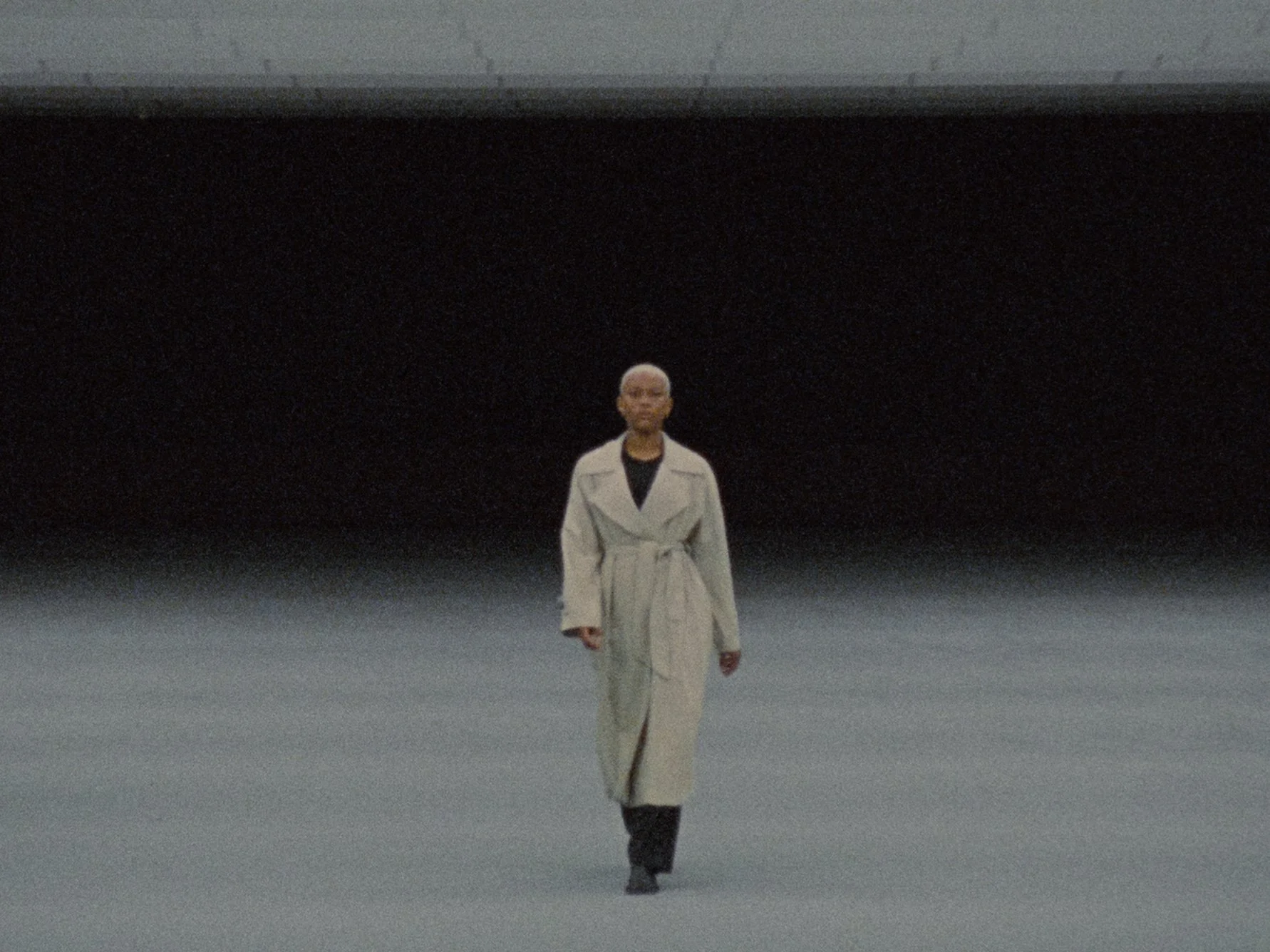 A woman with short white hair walking on a runway, wearing a long, light-colored trench coat and black boots. The background is dark and plain.