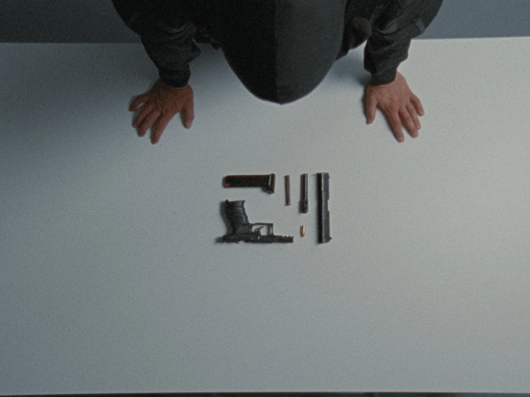 A person wearing black clothing is kneeling at a white table with various firearm parts and tools arranged on it, including a pistol grip, slide, barrel, and other gun components.