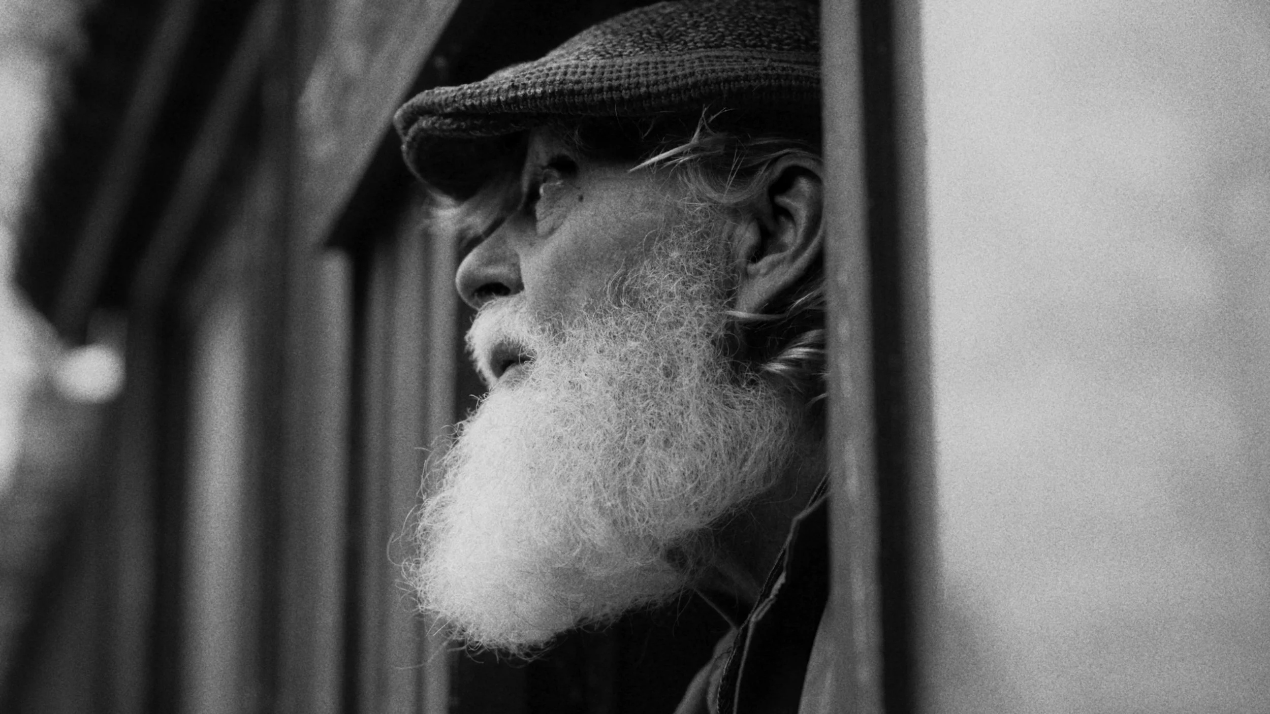 A black and white photo of an elderly man with a white beard, wearing a cap, looking to the side through a window or mirror.