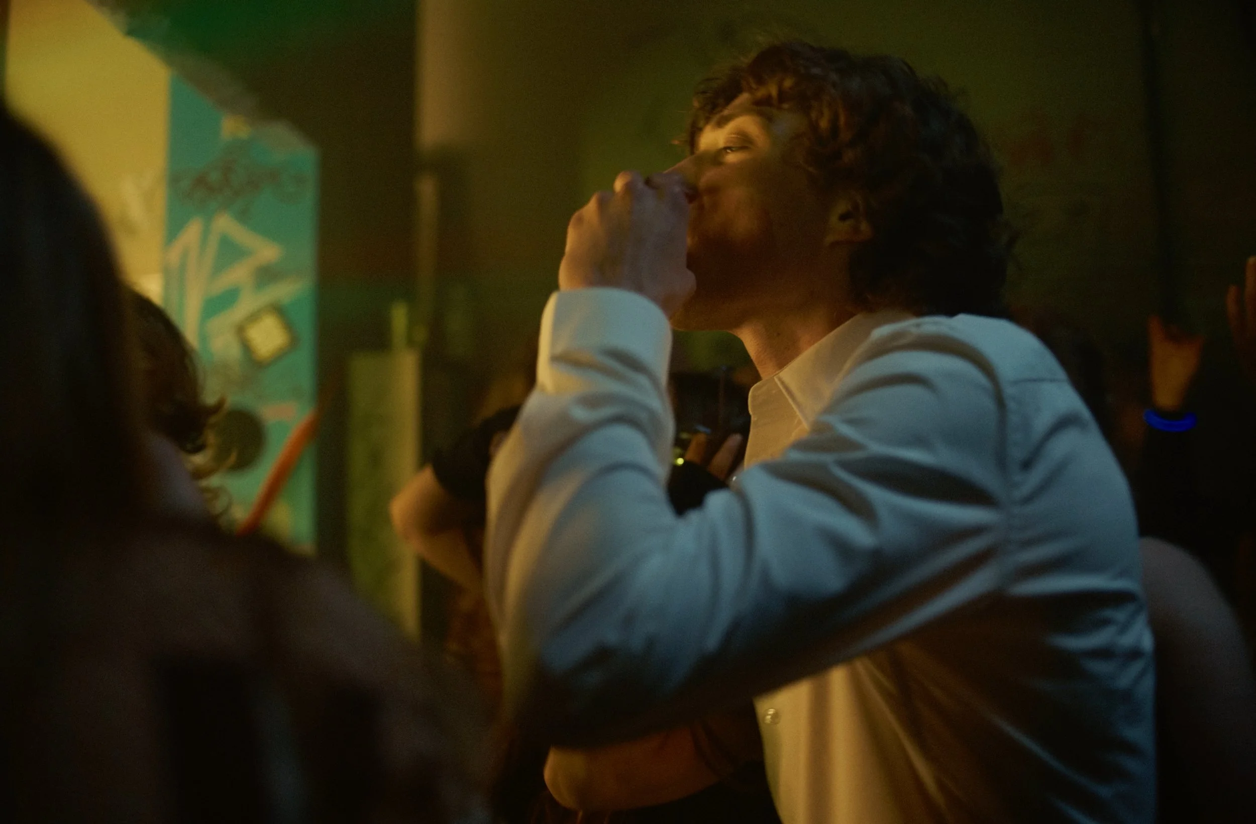 A person with curly hair wearing a white shirt is talking on a cell phone in a dimly lit setting, possibly a bar or club, with other people and colorful lights in the background.