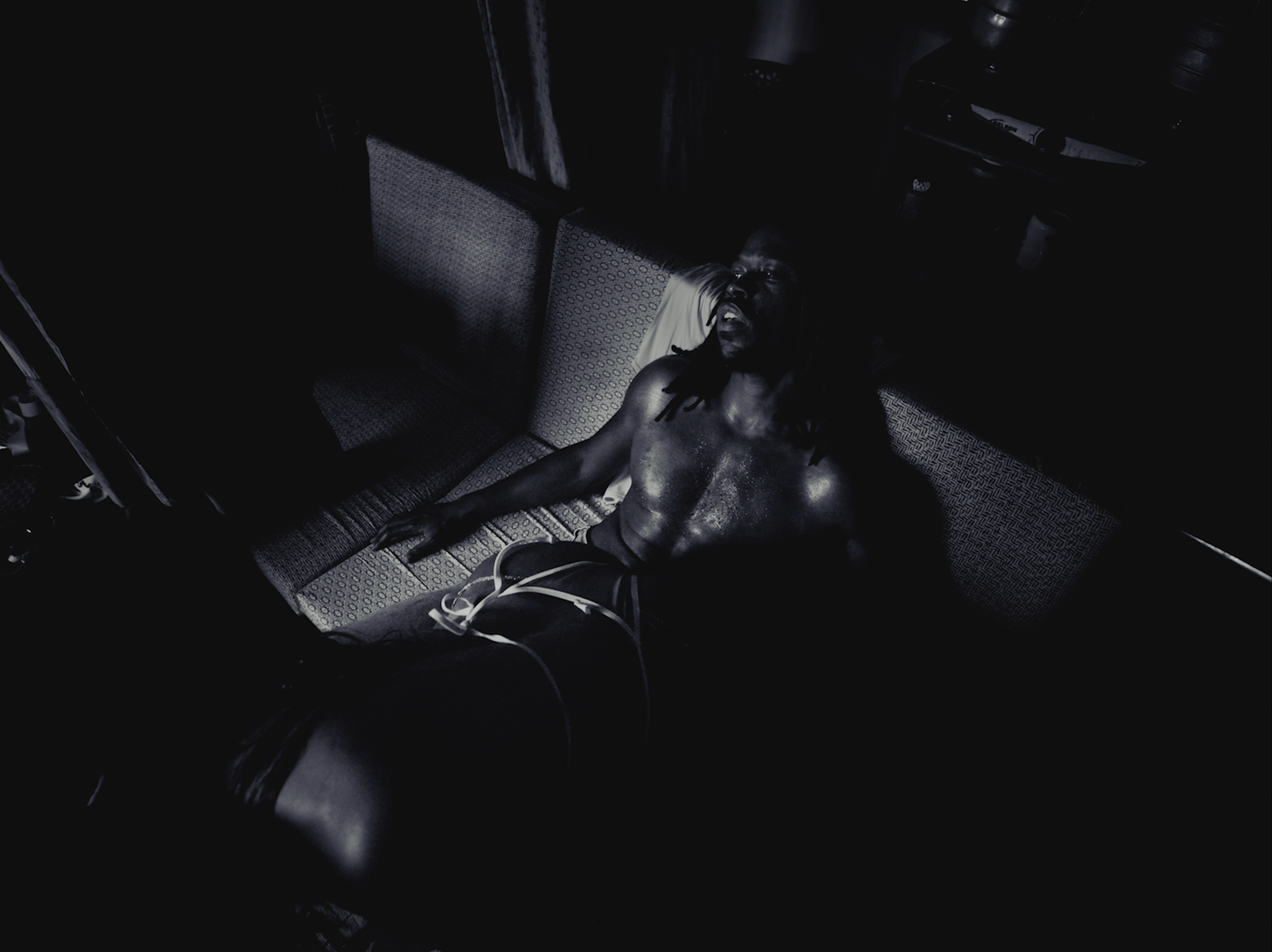A man with long dreadlocks and bare chest lying on a patterned couch in a dimly lit room.