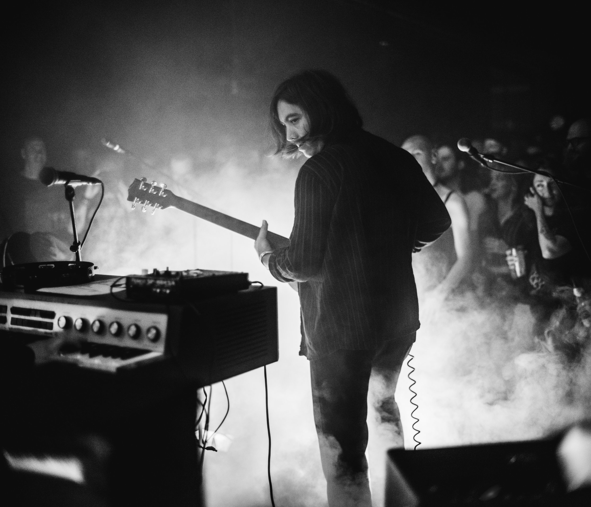 Black and white photo of a woman with shoulder-length hair playing an electric guitar on stage, with a crowd and microphones in the background.