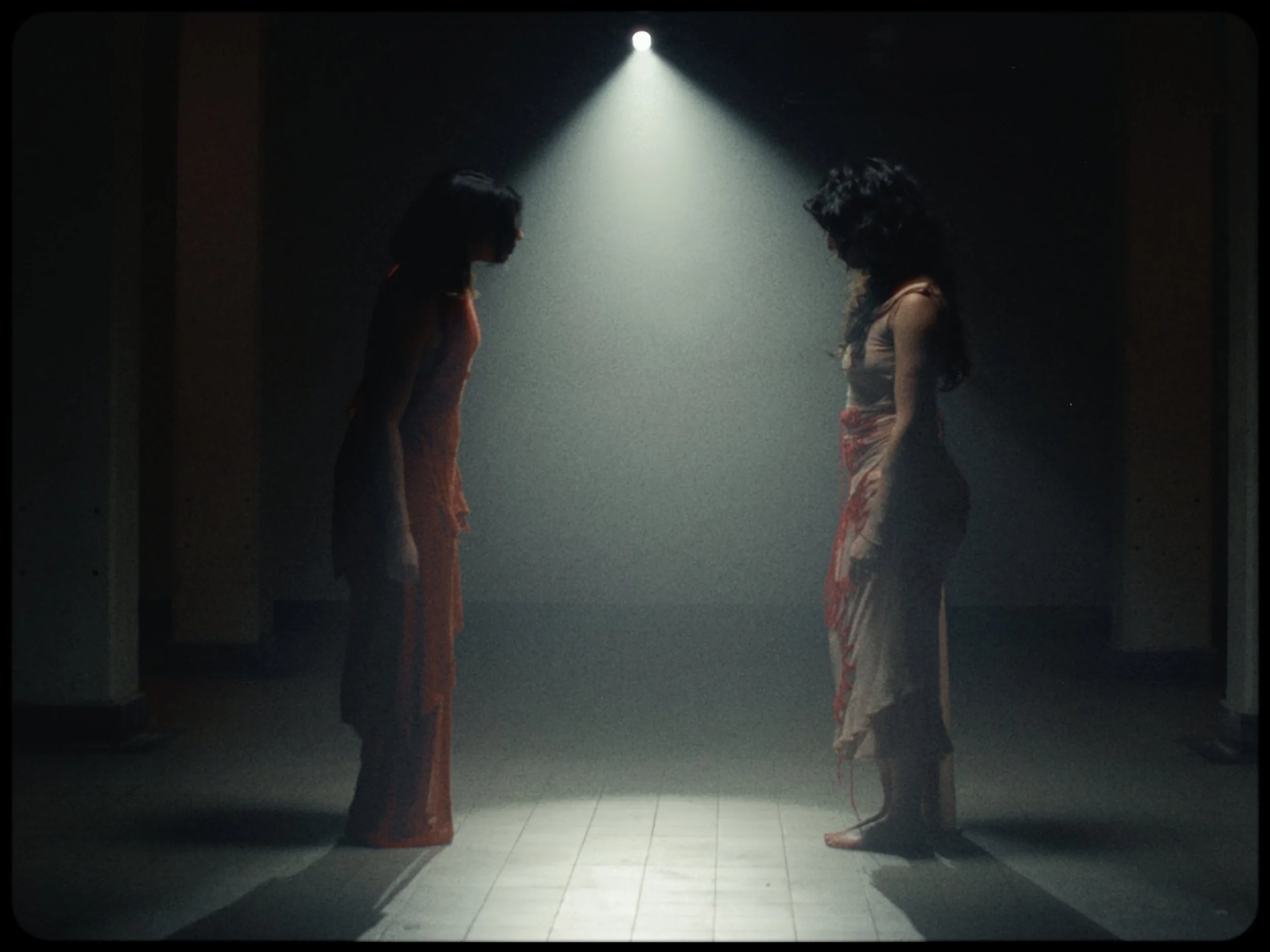 Two women with long dark hair standing face-to-face in a dark room illuminated by a single overhead light, dressed in colorful skirts and tank tops.