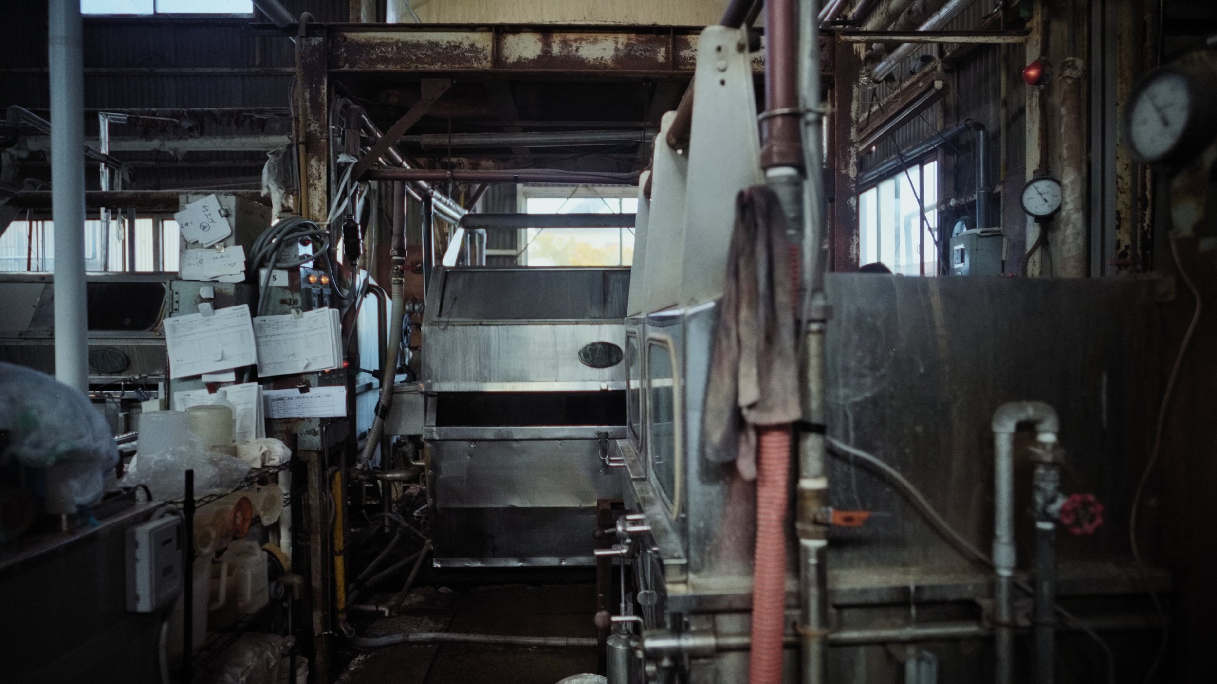Interior of an industrial kitchen or factory with stainless steel equipment, pipes, gauges, and handwritten notes on paper attached to machinery.
