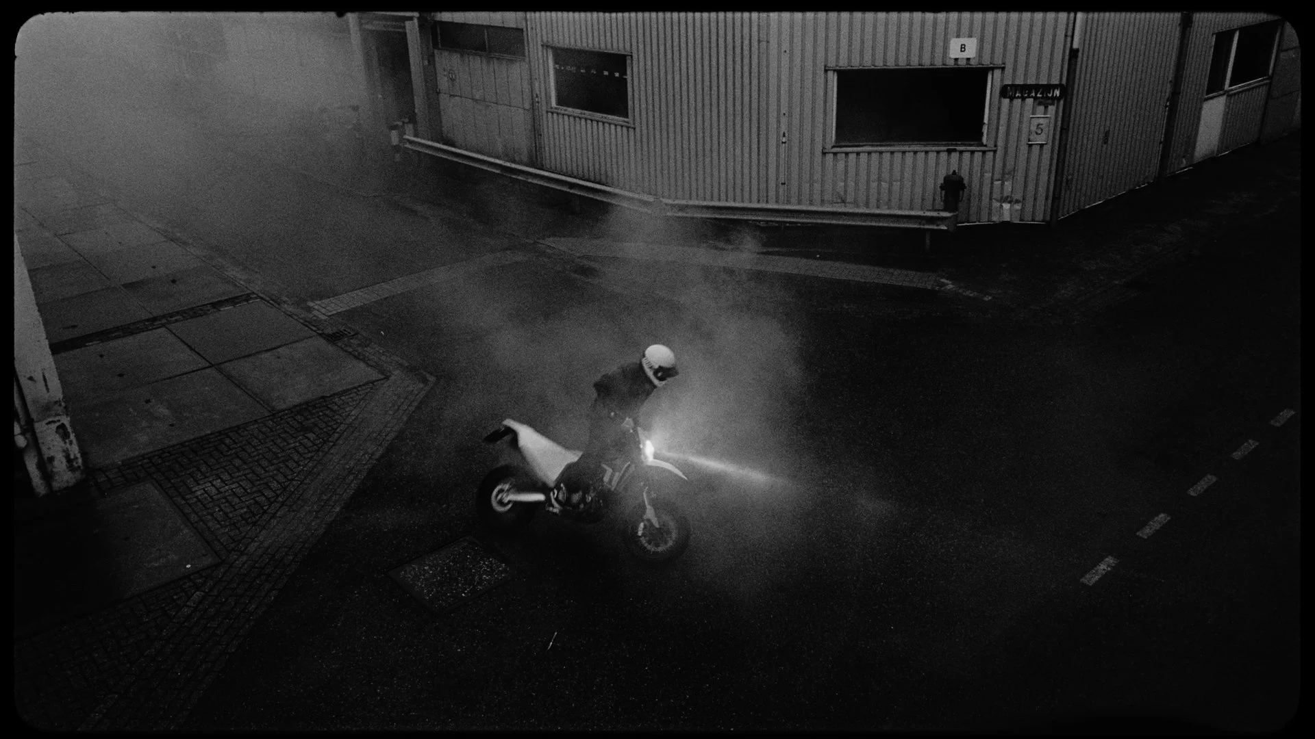 Nighttime street scene with a person on a motorcycle wearing a helmet, moving through water on the road. A building with windows and doors is visible in the background, with the street appearing wet.