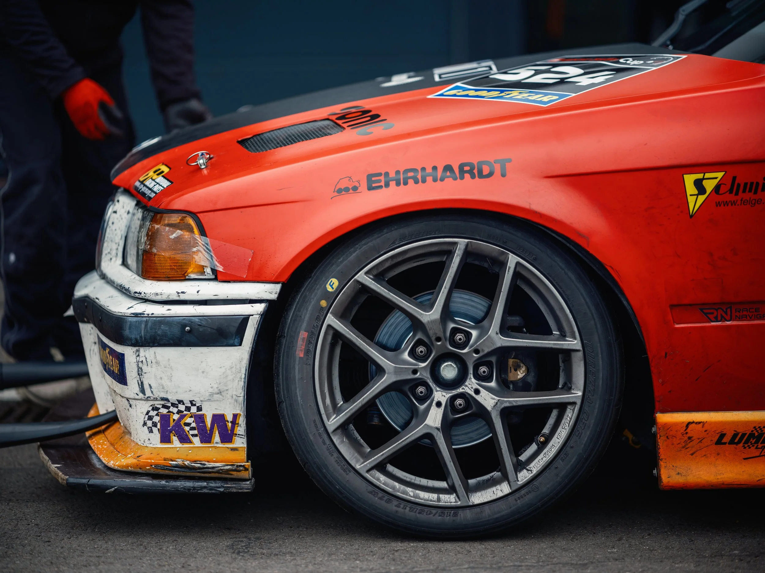 Close-up of a race car's front corner showing the wheel, tire, and part of the red body with sponsor decals, including 'EHRHARDT', 'KW', and others, along with a black vent on the hood.