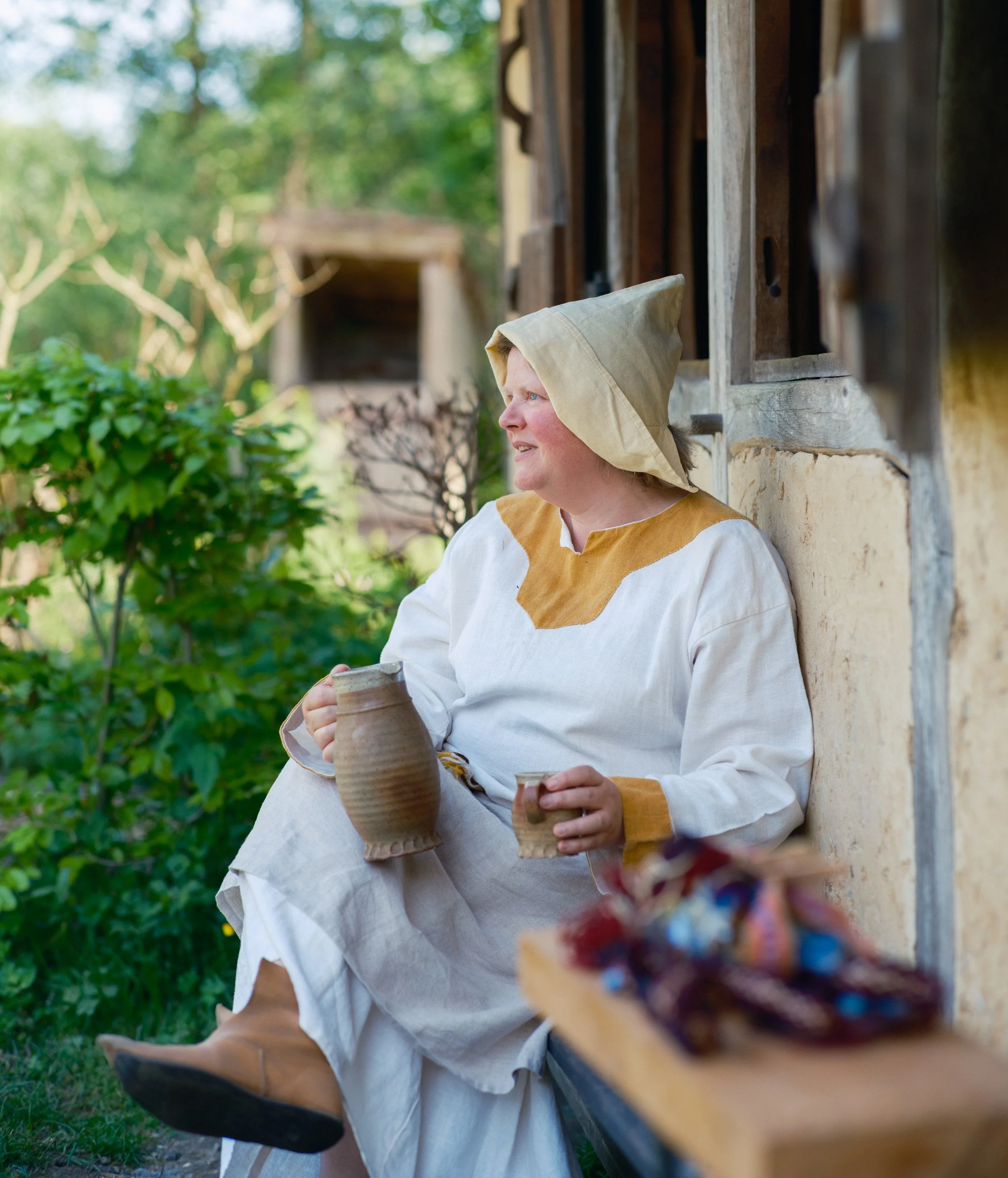 Woman in medieval-style clothing and a hooded cape sitting outdoors with a clay pitcher in her hands, near a wooden structure, with green trees and plants in the background.