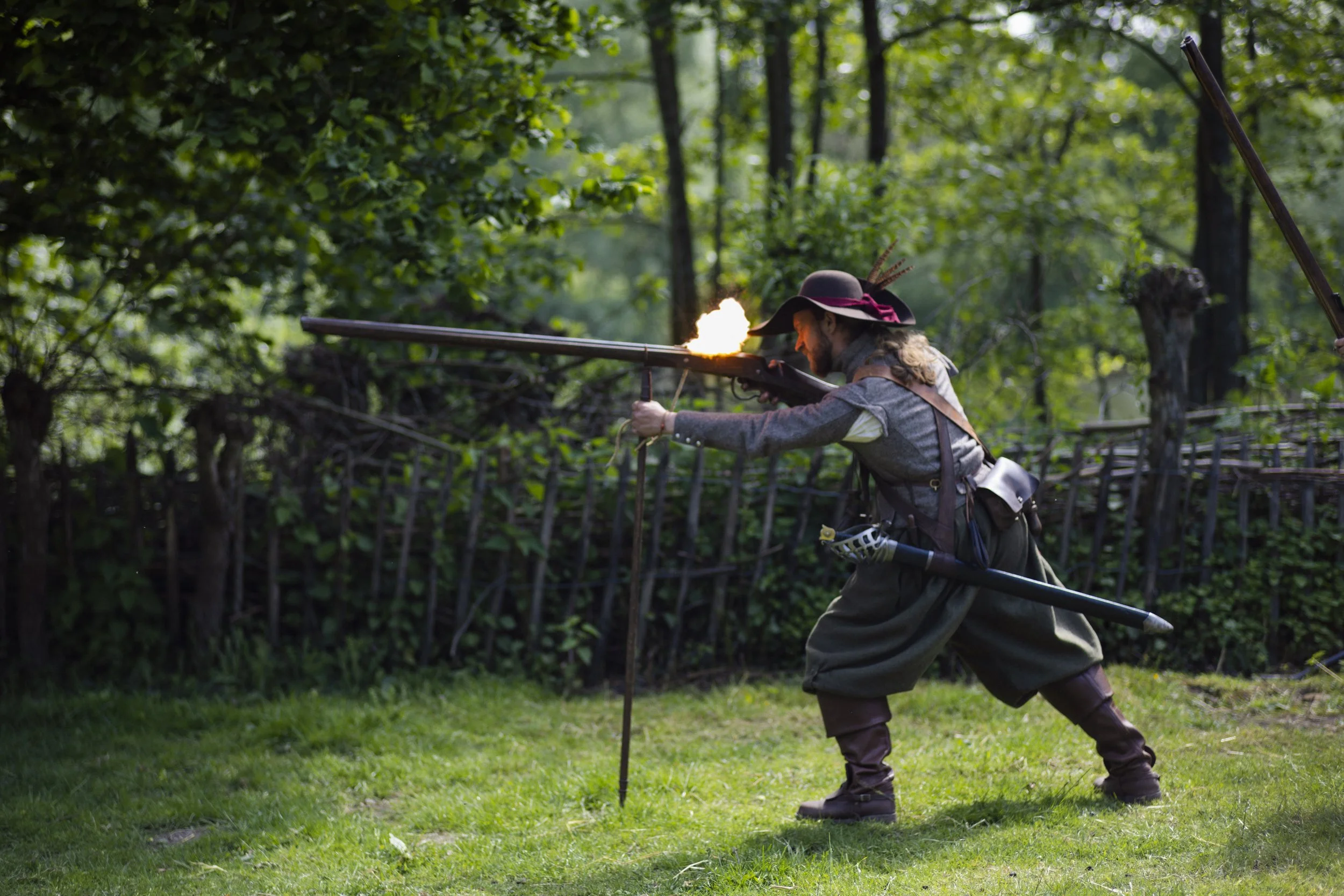A person dressed in Renaissance-style clothing, wearing a wide-brimmed hat with feathers, aiming a long, antique musket with a muzzle flash in a forested area. They appear to be participating in a historical reenactment or costume event.