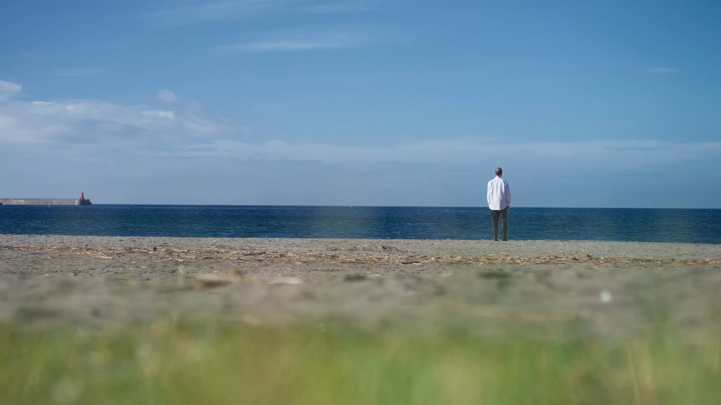 A man standing alone on a sandy beach facing the ocean with a lighthouse in the distance under a partly cloudy sky.