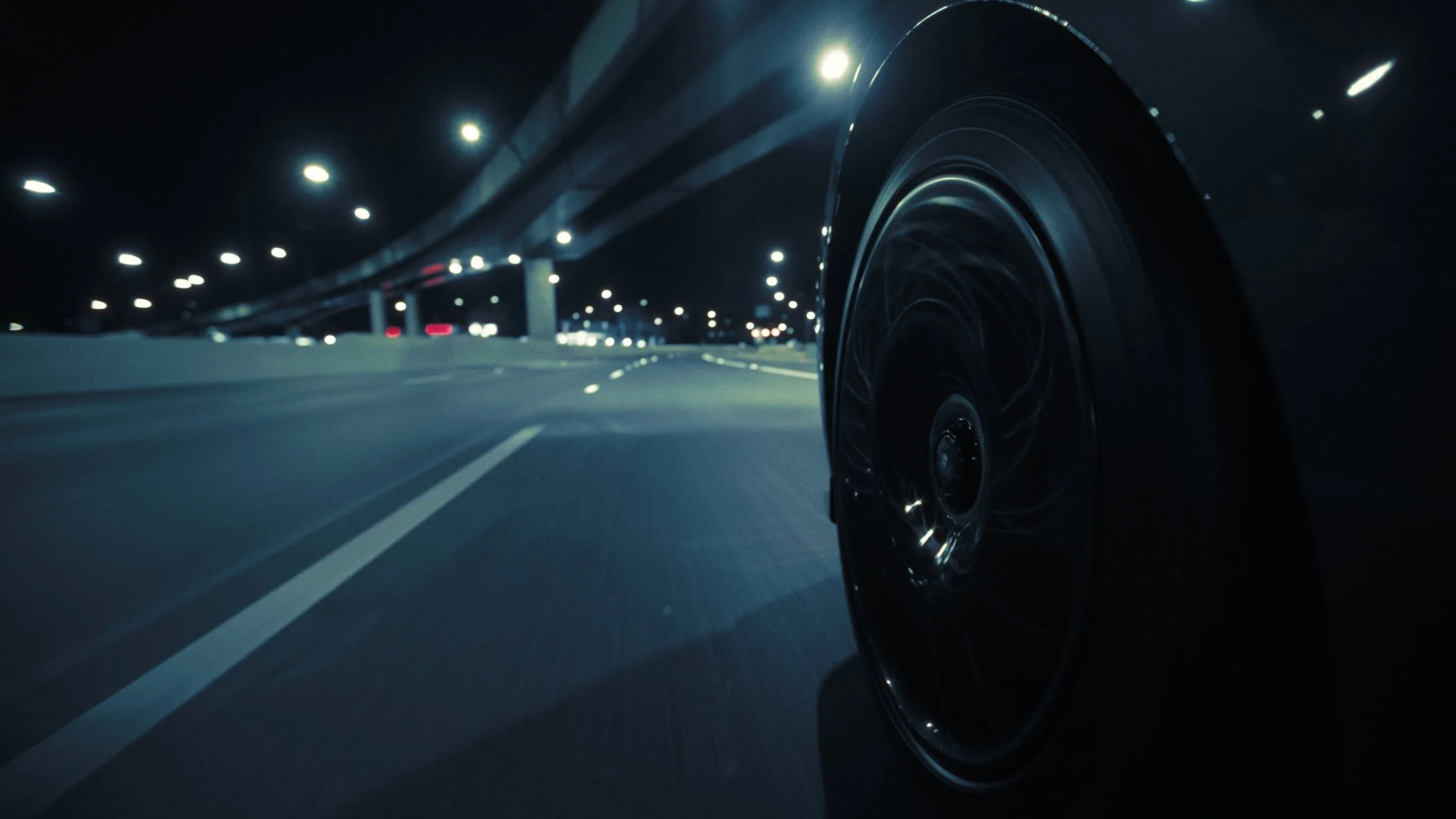 Nighttime view of a car driving on a highway, with a close-up of the wheel on the right side and streetlights illuminating the road.
