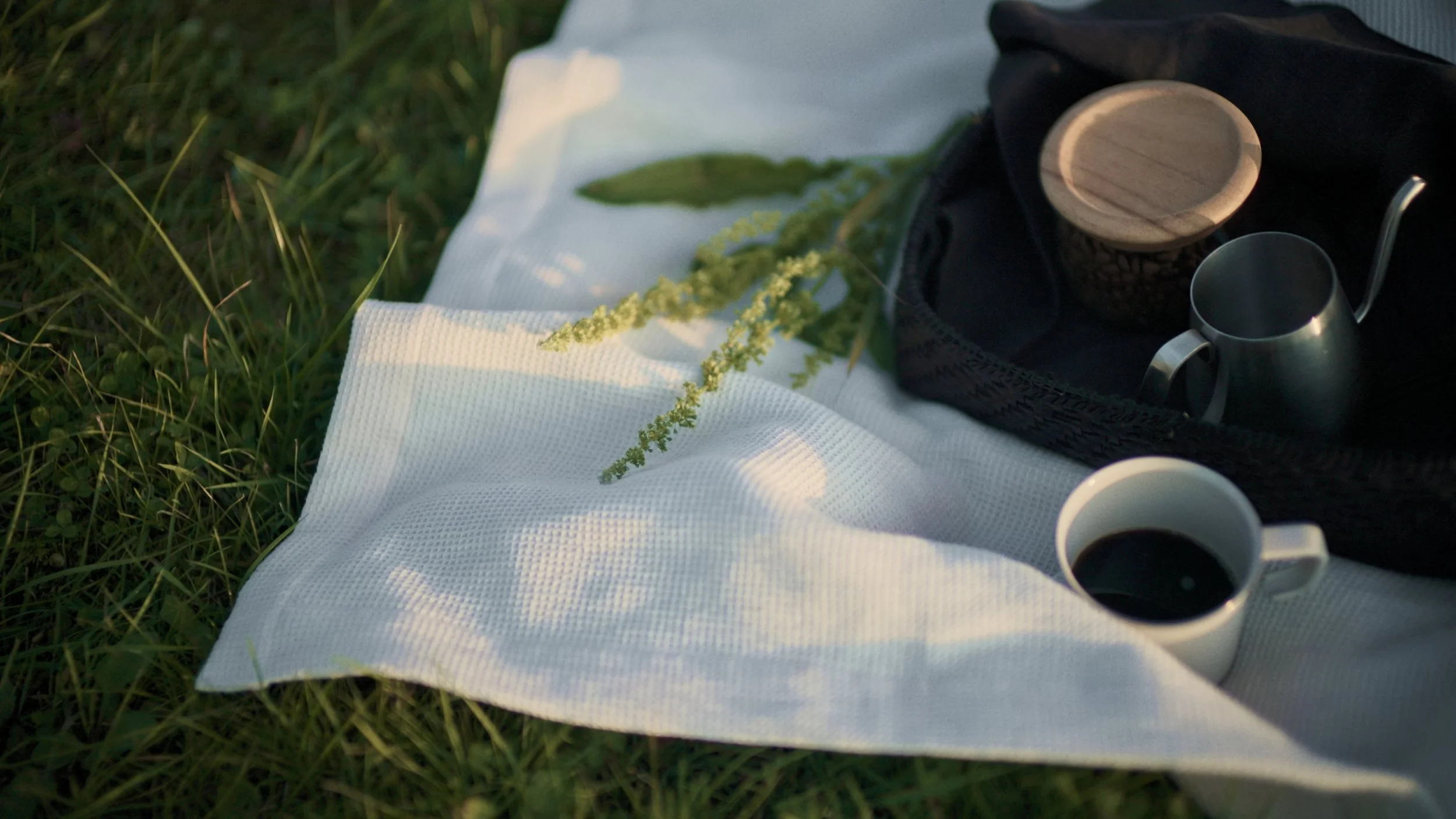 A picnic setup on grass including a white cloth, a cup of coffee, a metal mug, a black bag with a wooden container and a metal cup inside, and some green plants.