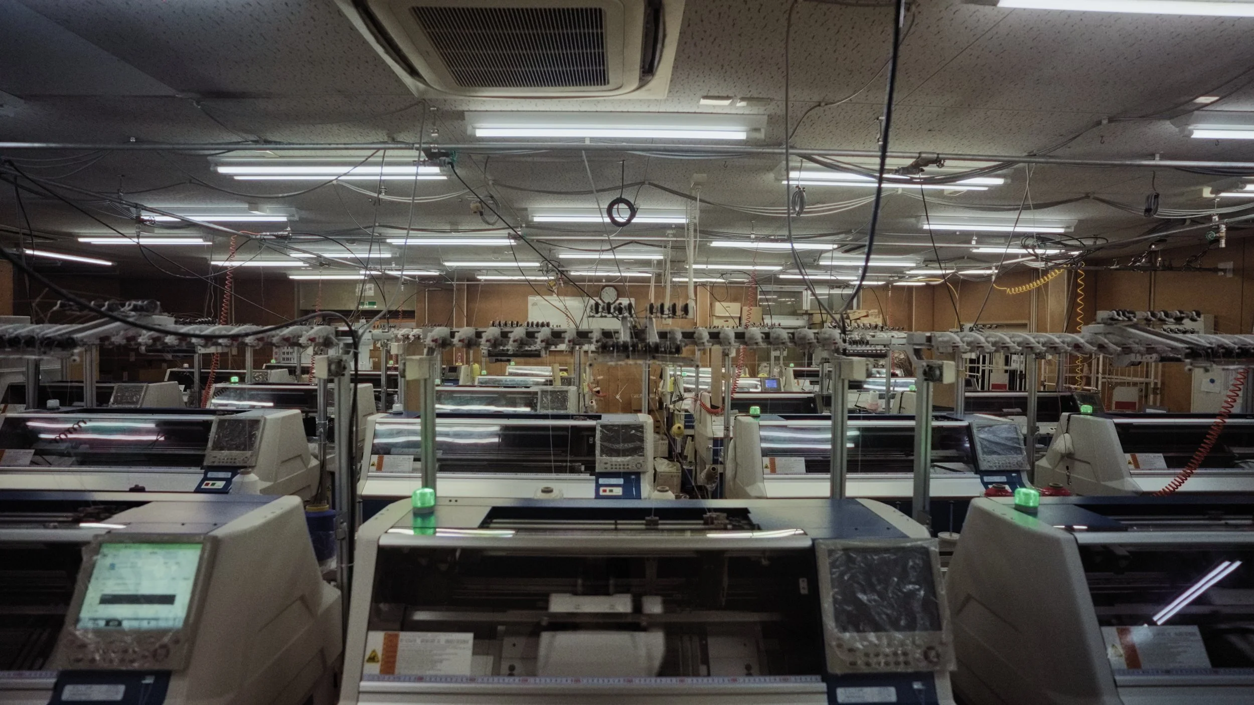 A room filled with multiple electronic machines and monitors, with numerous overhead fluorescent lights and an intricate network of wires hanging from the ceiling.