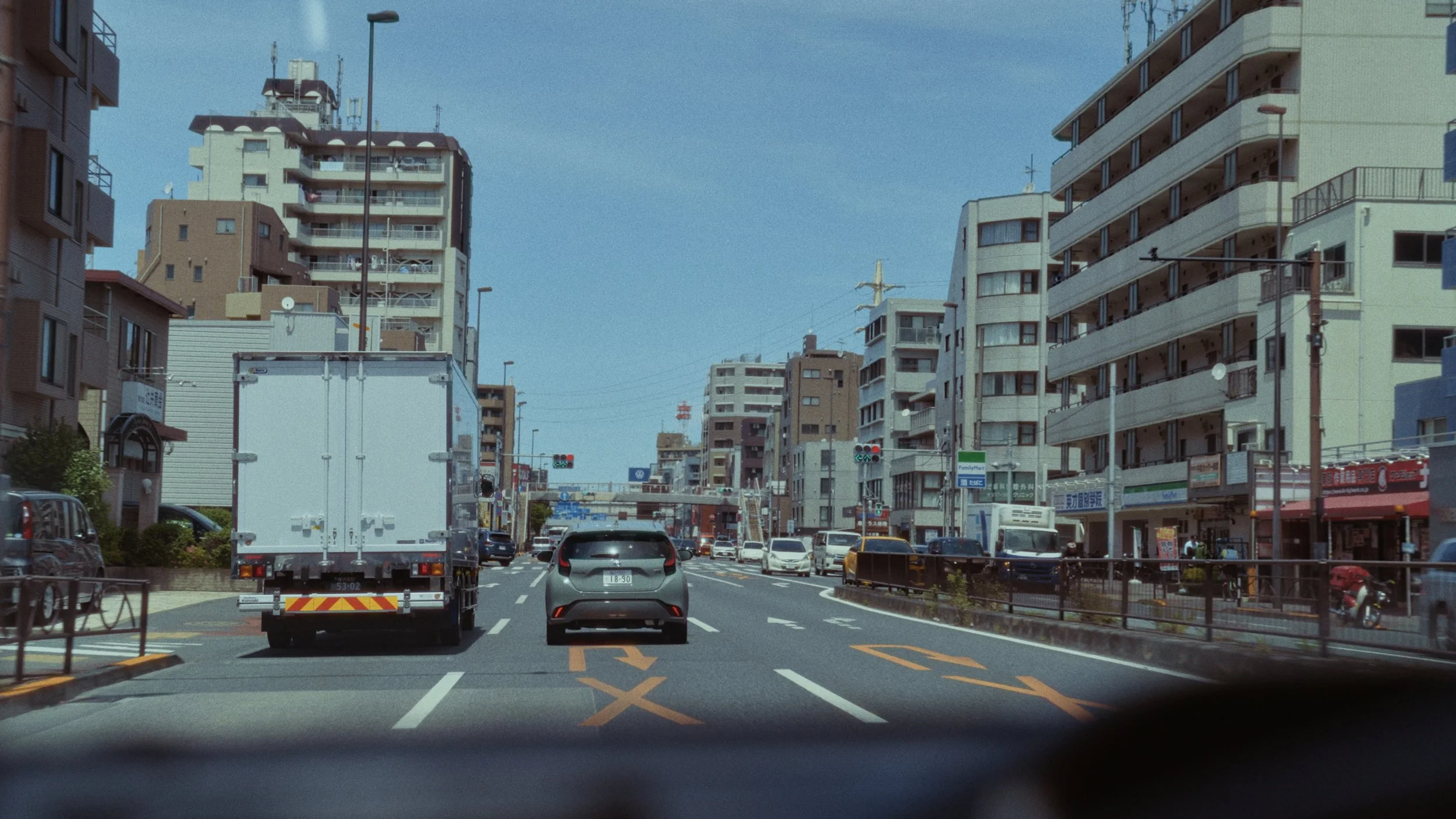 A city street scene with multiple tall buildings on each side, cars and trucks on the road, traffic signals, and a blue sky with a few clouds.
