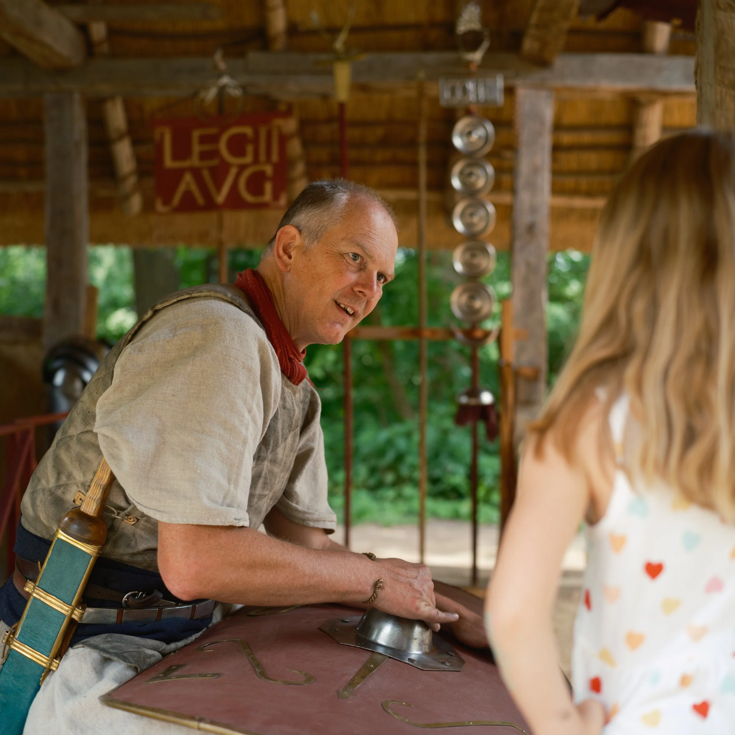 A man in historical costume playing a musical instrument under a wooden structure with a red sign that reads 'LEGAL' and other metallic objects hanging in the background.