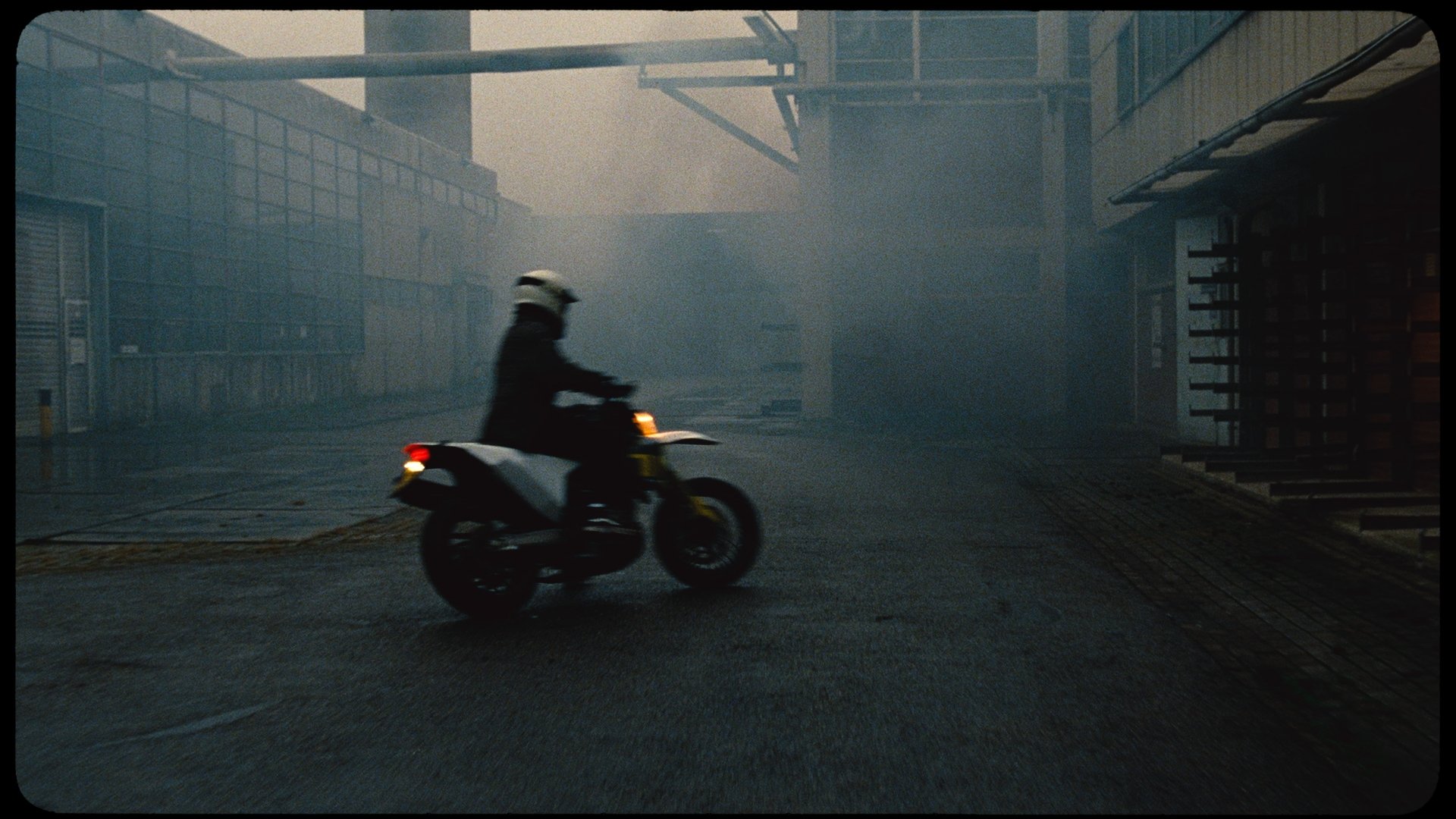 A motorcyclist wearing a helmet riding a motorcycle on a foggy, deserted urban street.