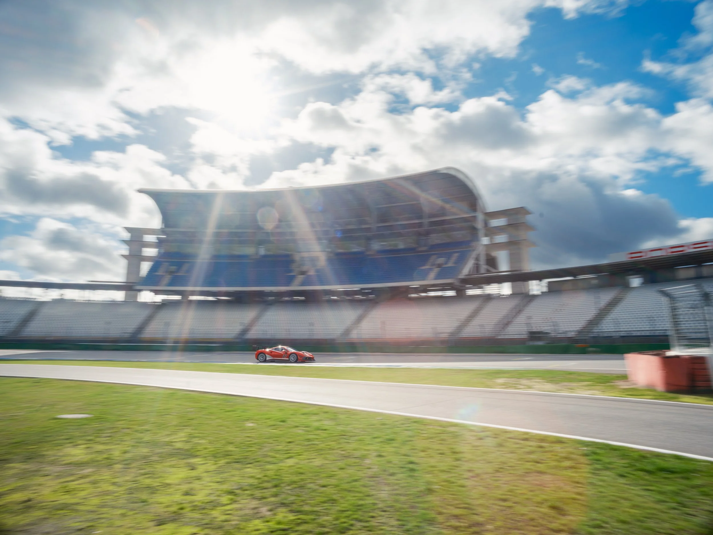 A race car driving on a track with a modern stadium and cloudy sky in the background.