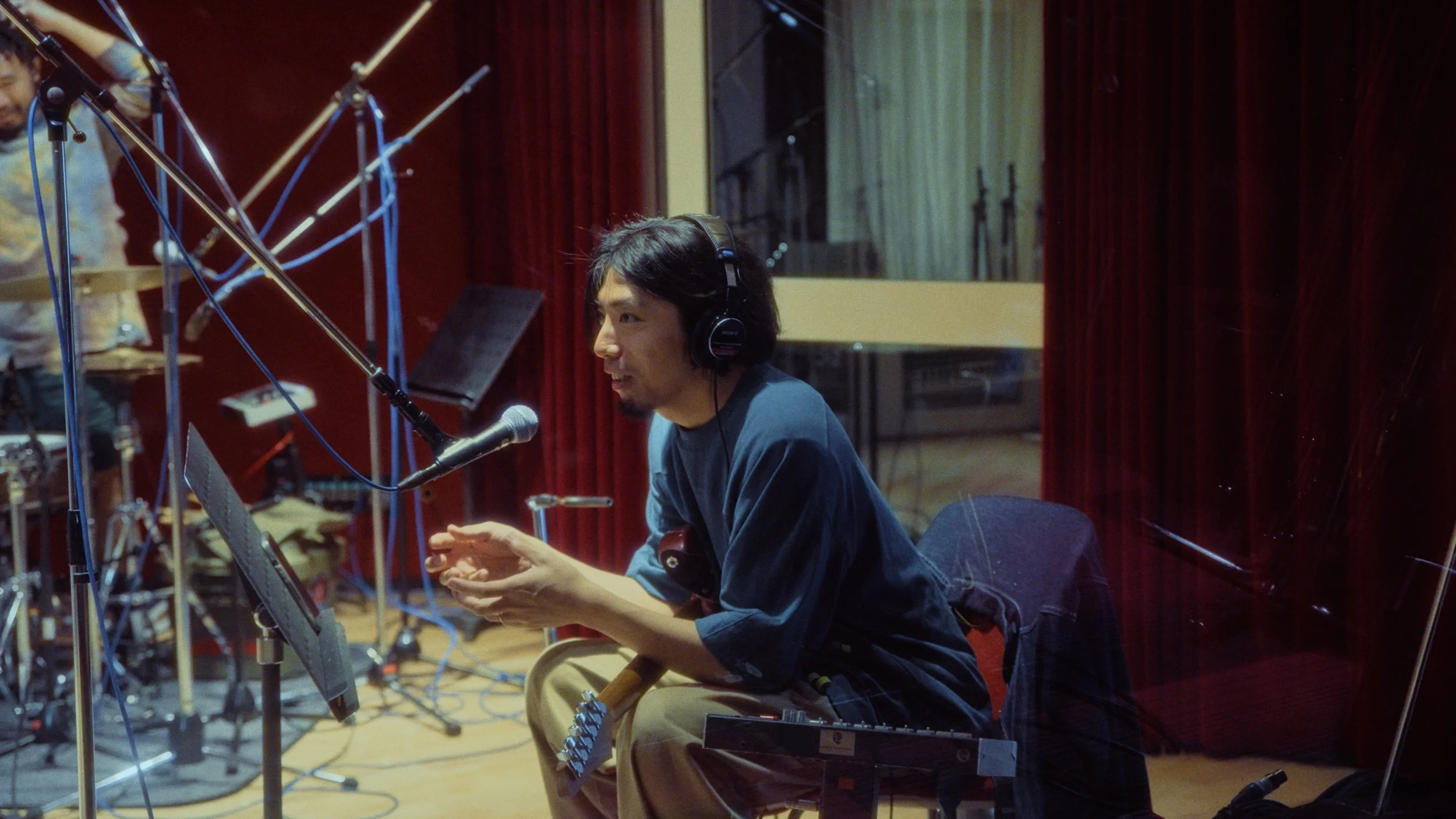 A man with dark hair and headphones sitting at a recording studio, speaking into a microphone, with music equipment and recording gear around him.