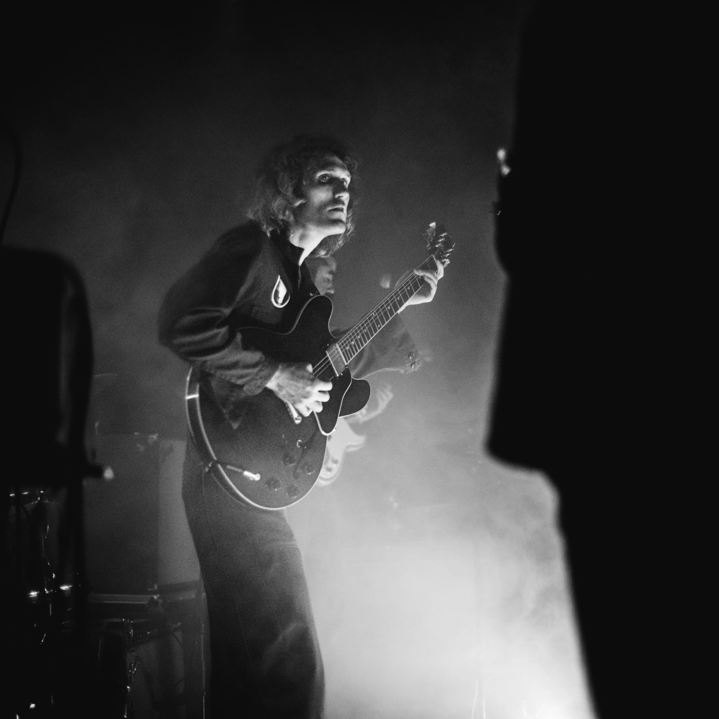 Black and white photo of a person playing a guitar, looking thoughtful or intense, illuminated against a dark background.