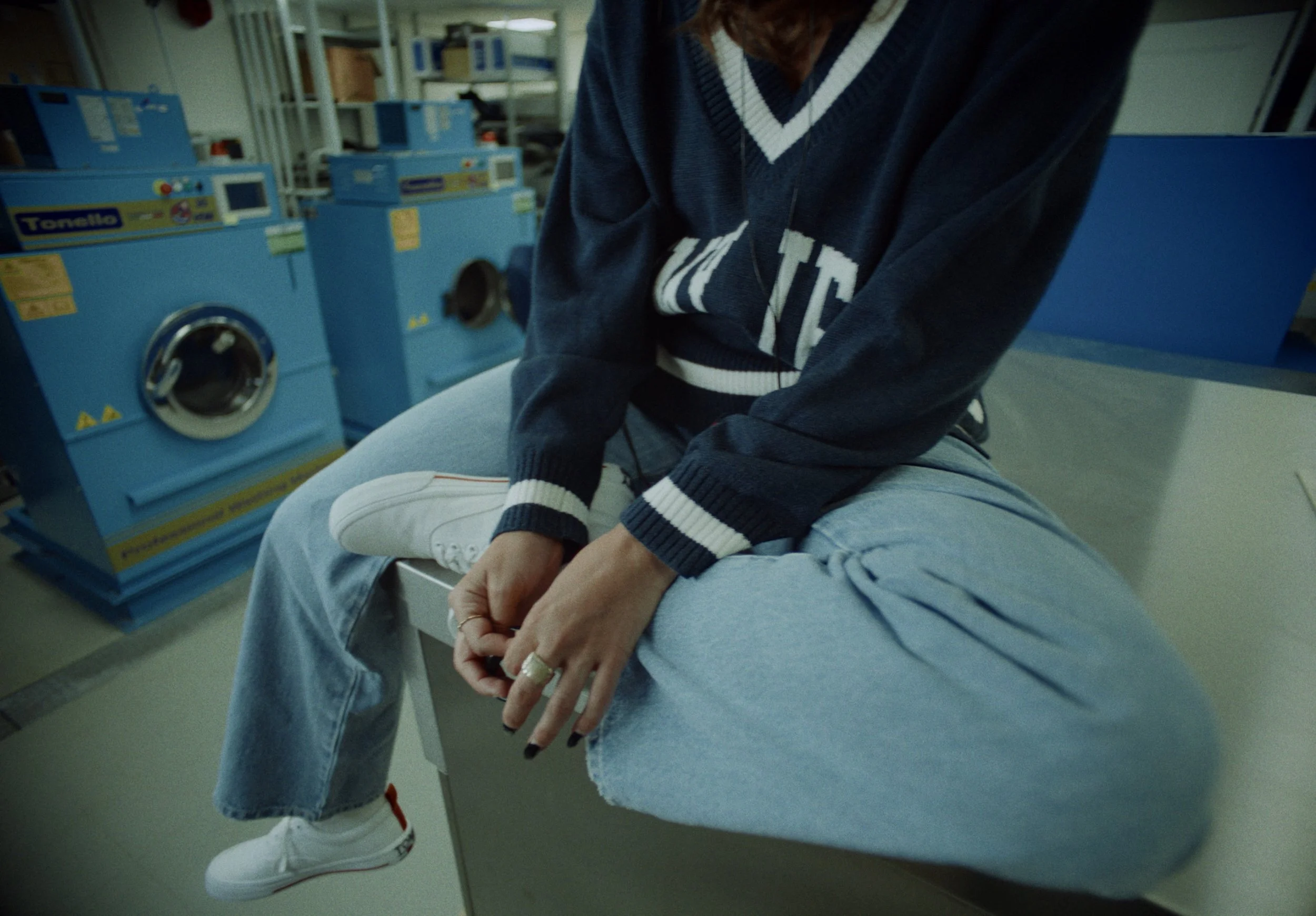 A person sitting on a laundry basket in a laundromat, wearing a blue sweatshirt, light blue jeans, and white sneakers, with laundry machines in the background.