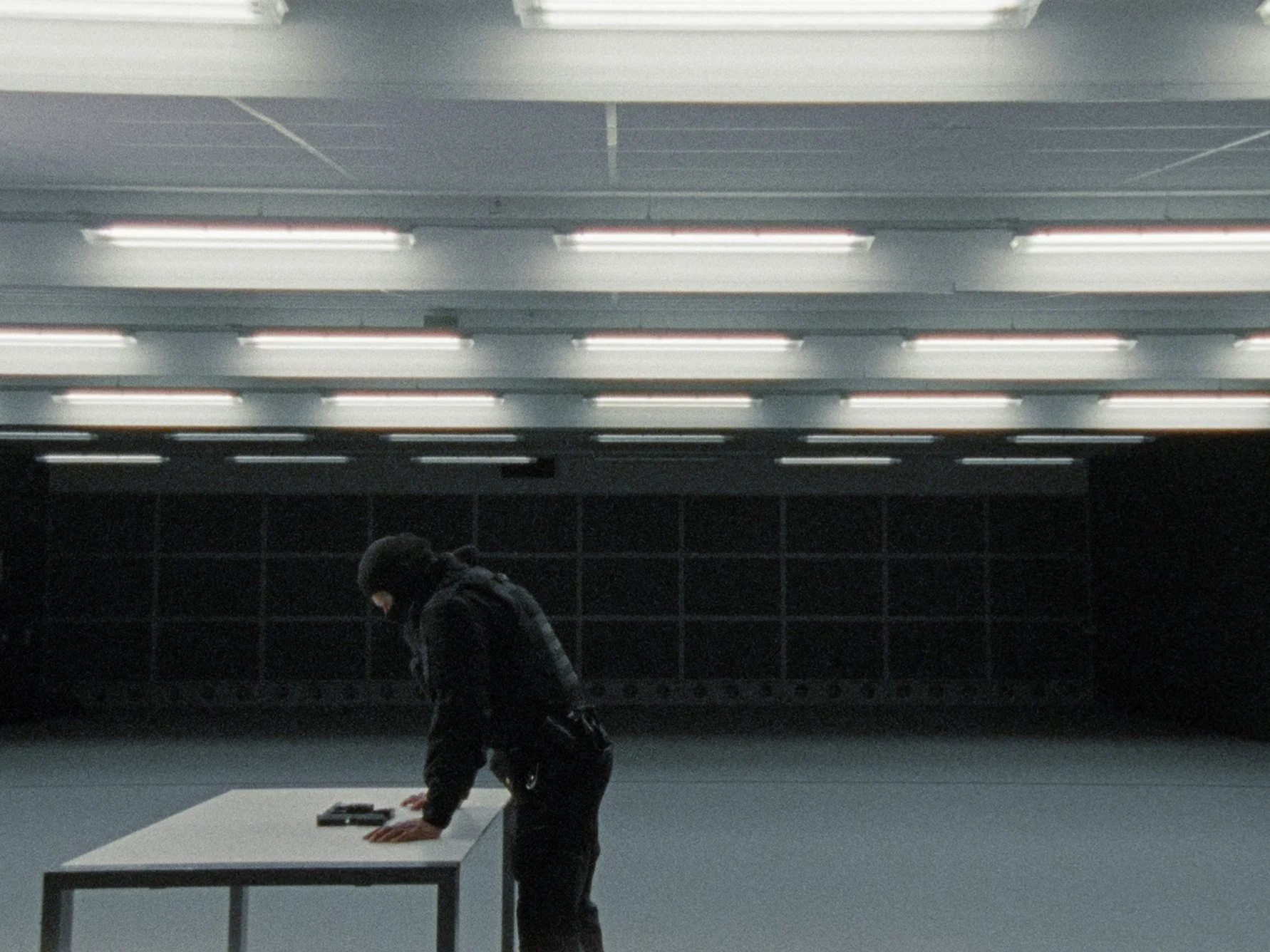 A person with dark hair and a dark coat leaning over a white table in a modern, underground space with fluorescent lights and black tiled walls.