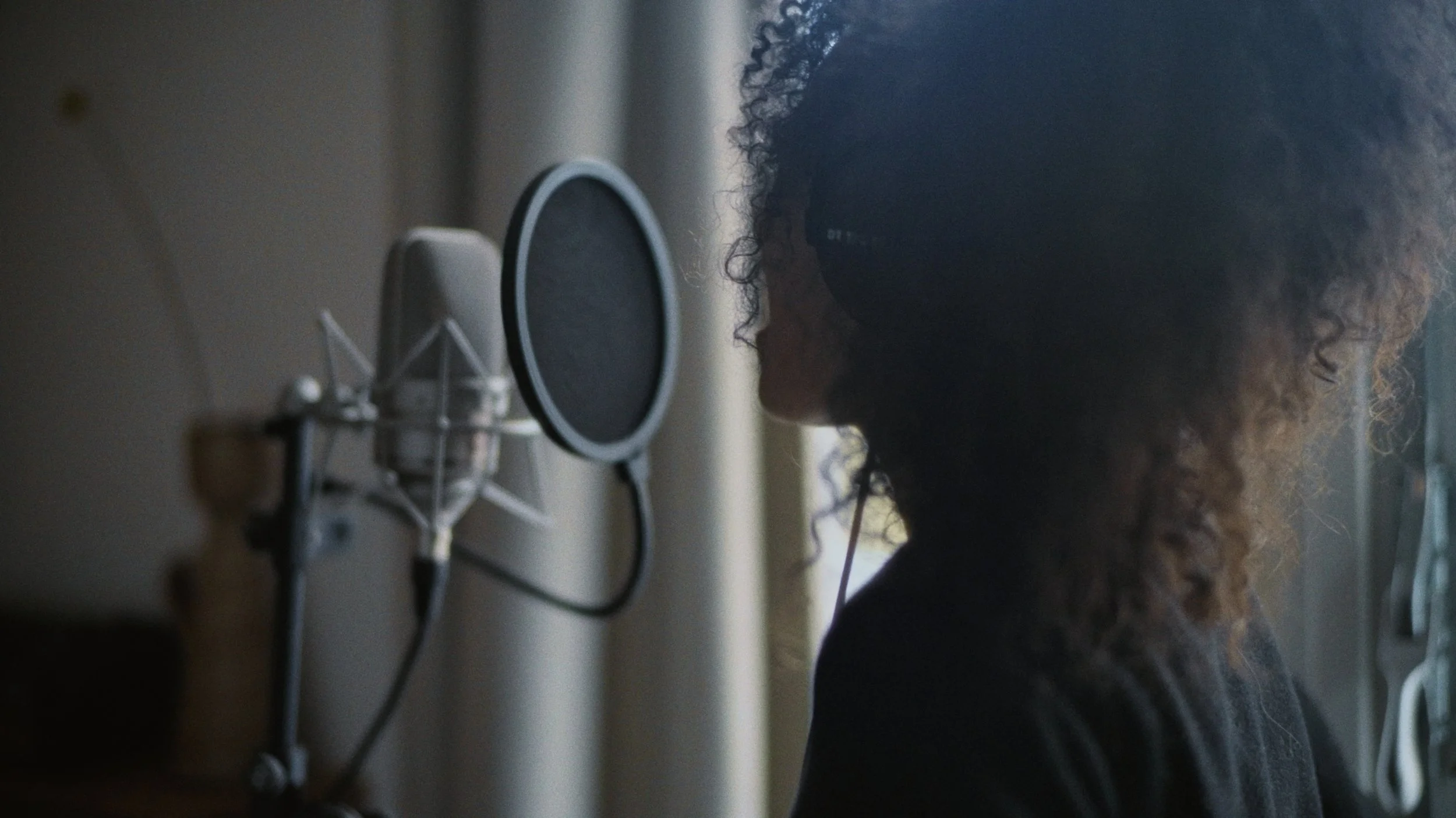 A person with curly hair recording in a soundproof room with a microphone and headphones.