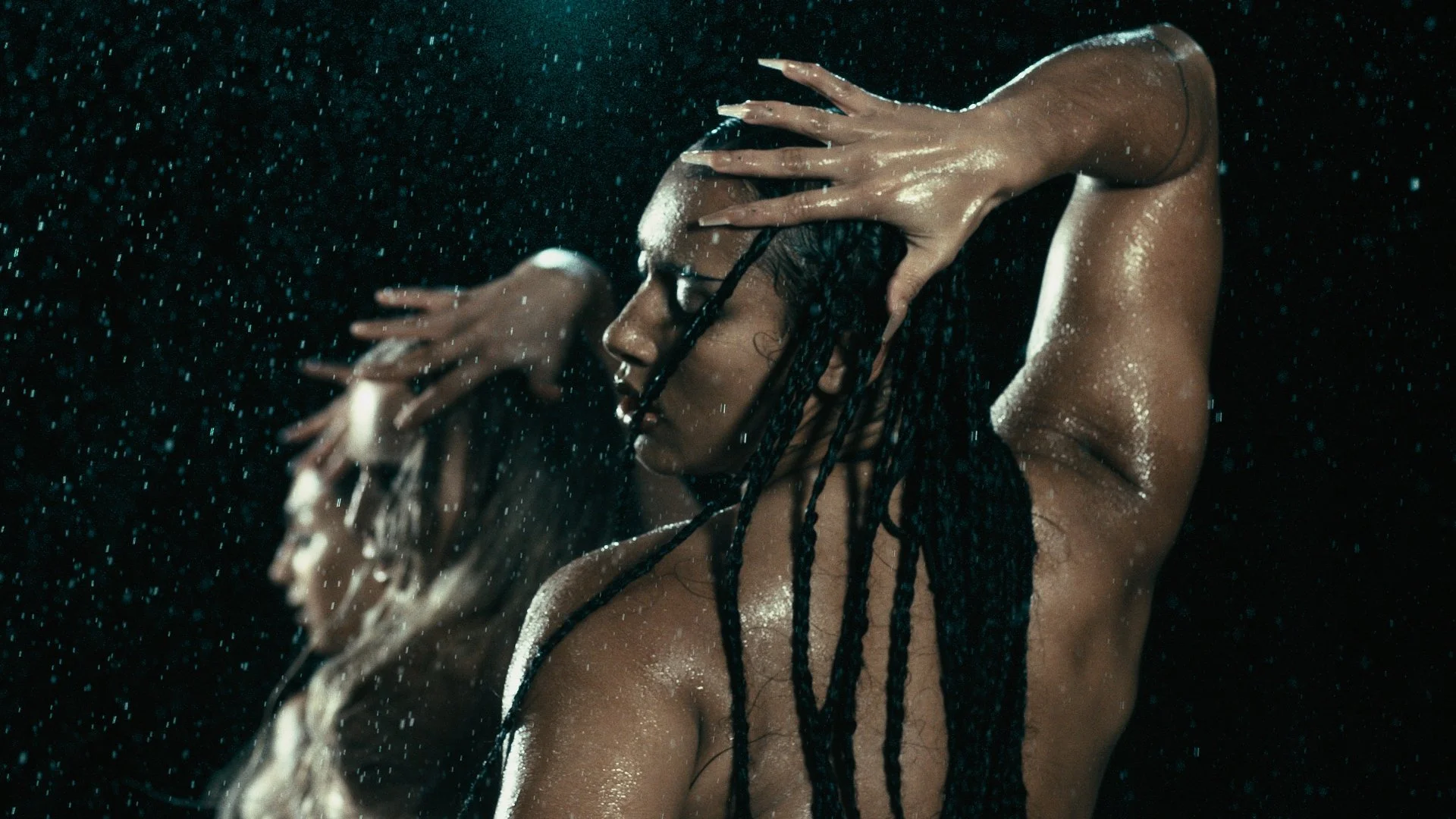 A woman with braided hair is under water with water droplets falling around her, eyes closed, with her arms raised and hands around her head.