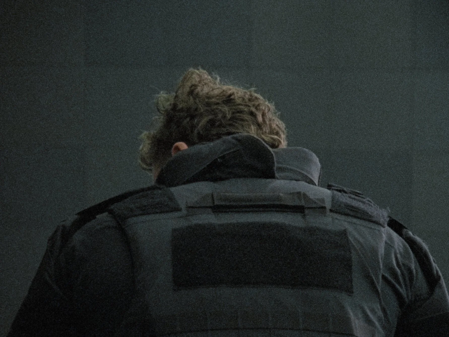 Back view of a person with curly hair wearing a dark jacket and jeans, standing in a dark environment.