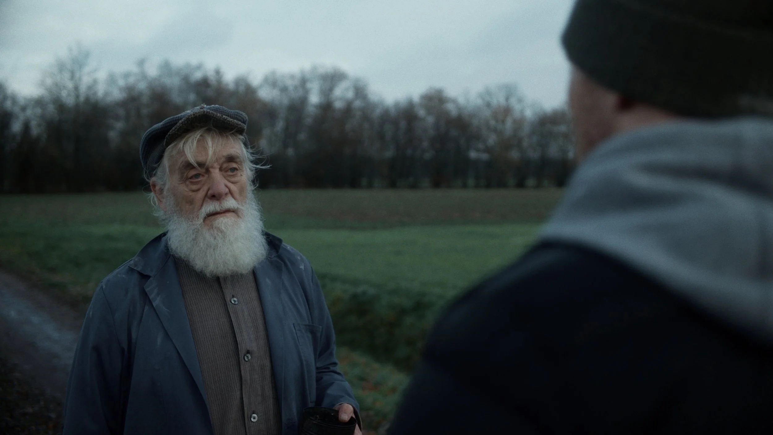 Older man with white beard and mustache, wearing a dark hat and jacket, looking at a younger man in a hoodie outdoors in a park or field with trees in the background.
