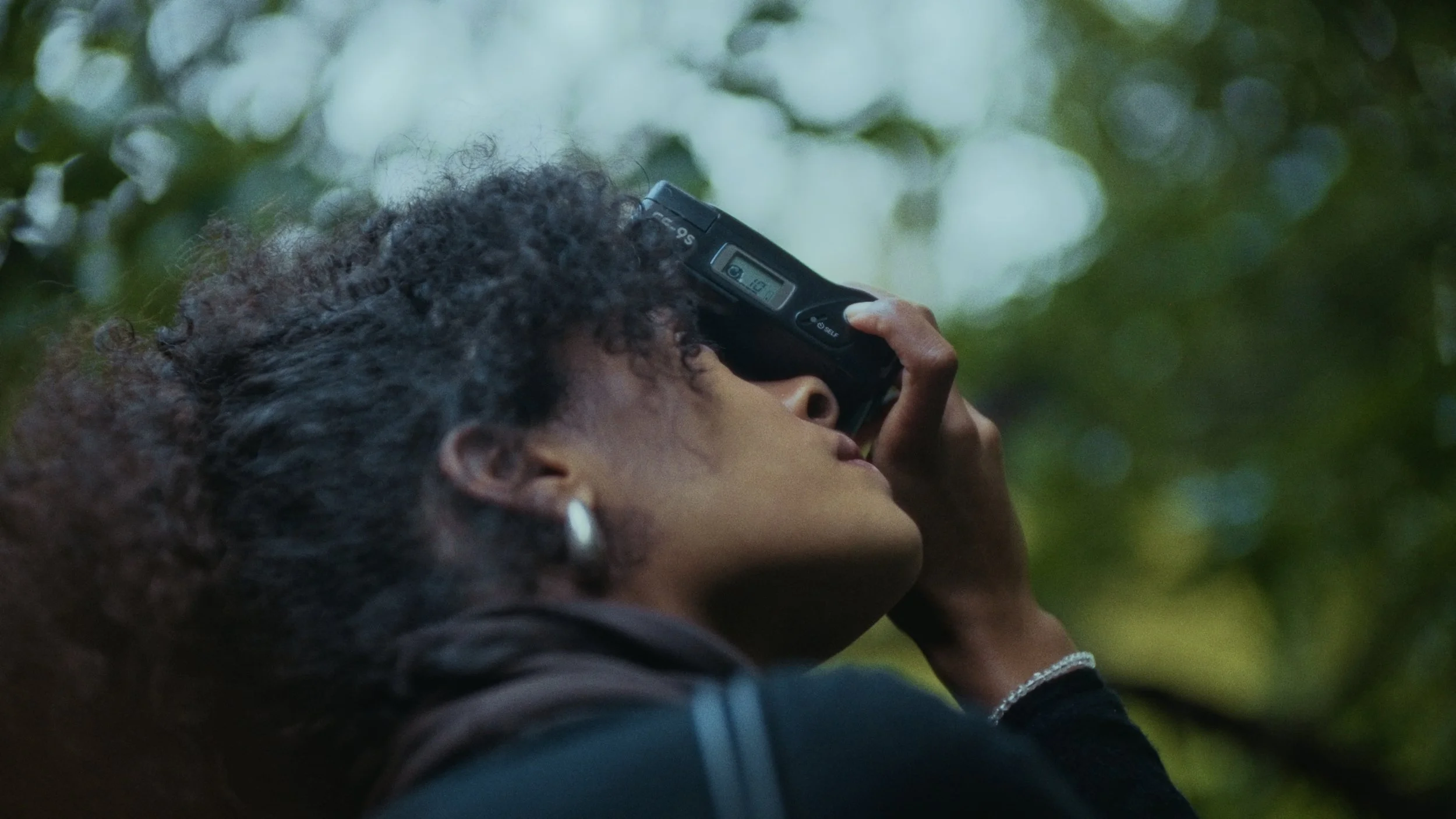 A person with curly dark hair and earrings is looking up at the sky through a viewing device in a green, leafy outdoor setting.