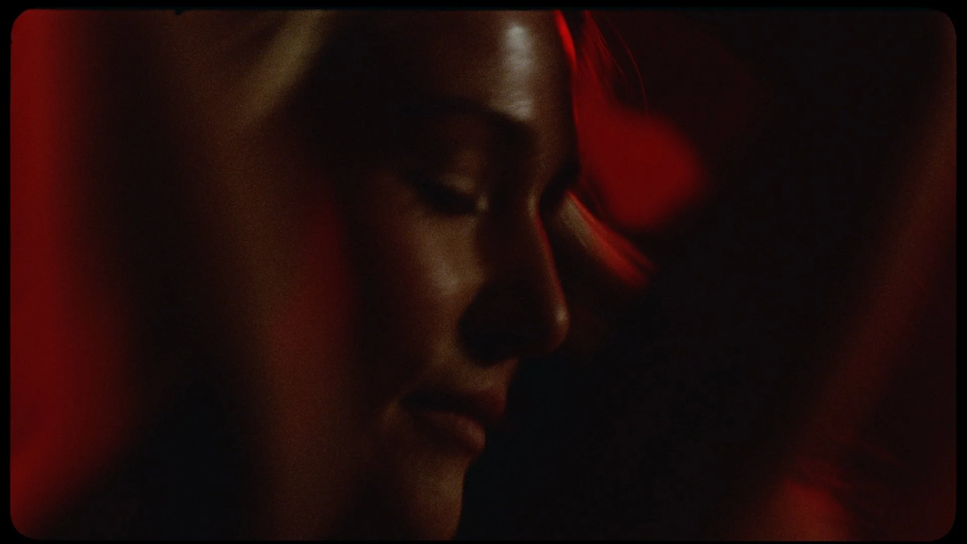 Close-up of a woman's face in profile, eyes closed, with red lighting highlighting her features, against a dark background.