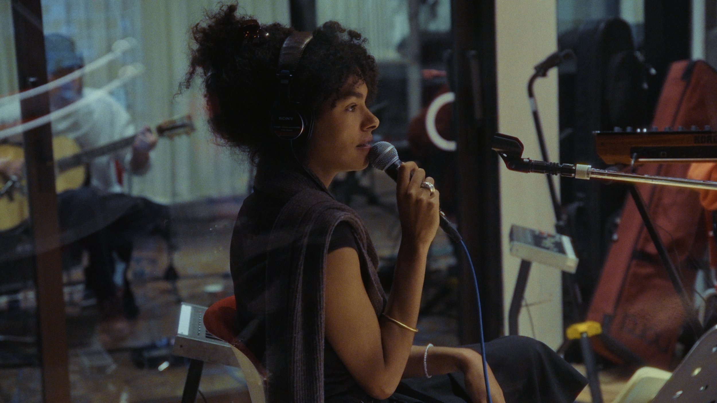 A woman with curly hair wearing headphones and a ring, holding a microphone, sitting in front of a musical keyboard in a recording studio.