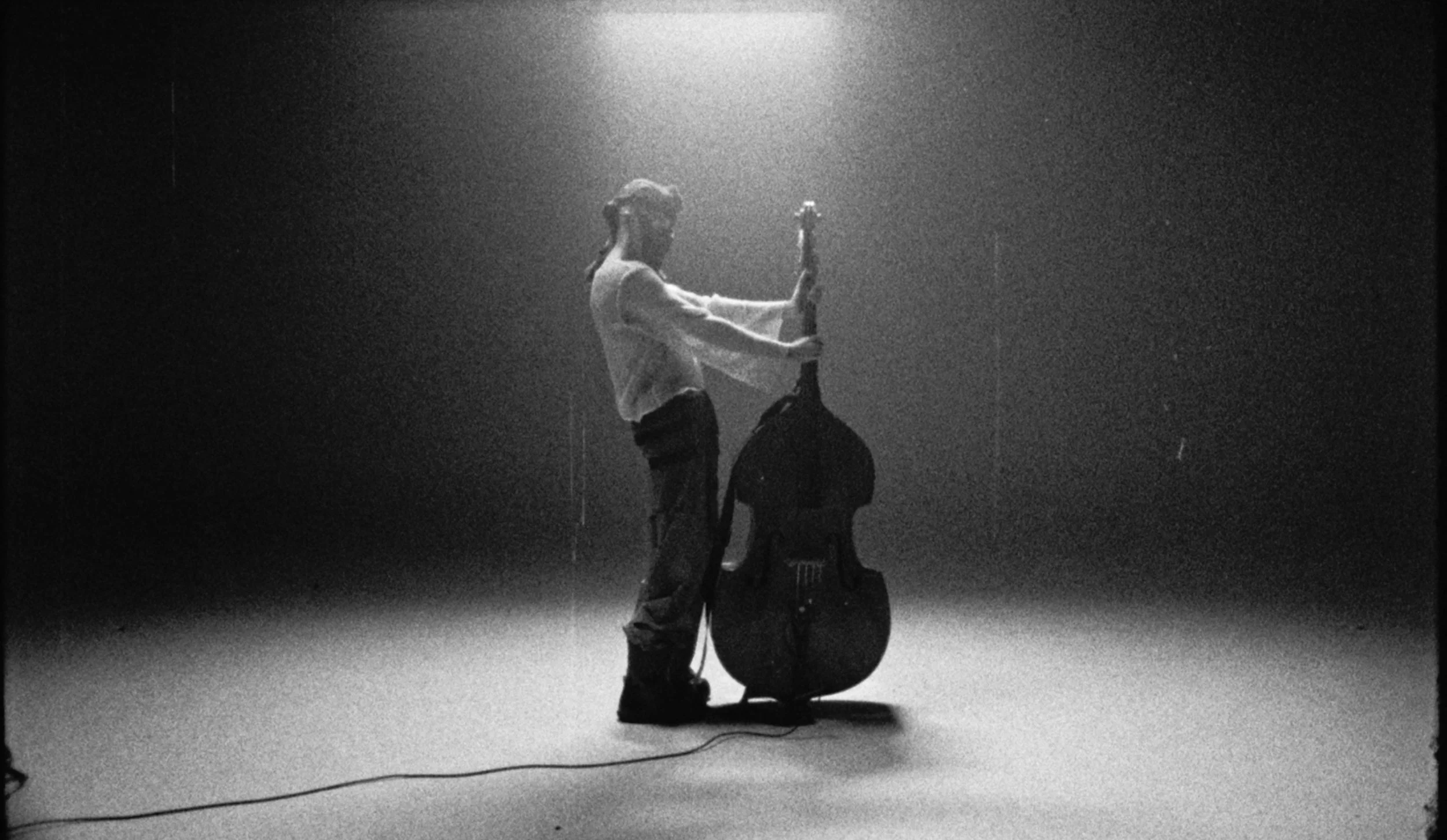 A woman with a ponytail standing in a dark space, holding a microphone and a guitar case, with a backpack on the floor.