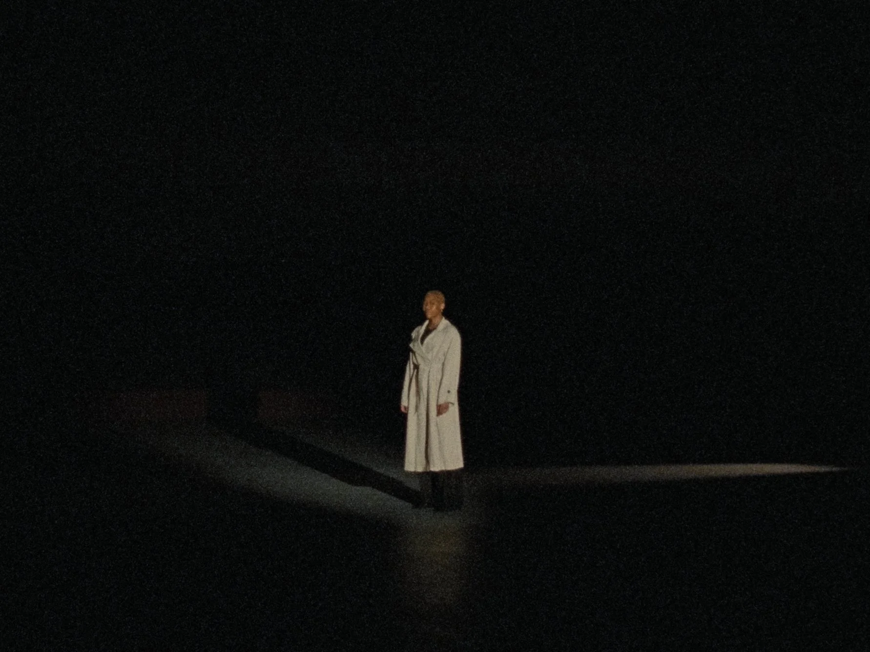 A person standing alone on a dark stage, wearing a long light-colored coat, with a plain black background.