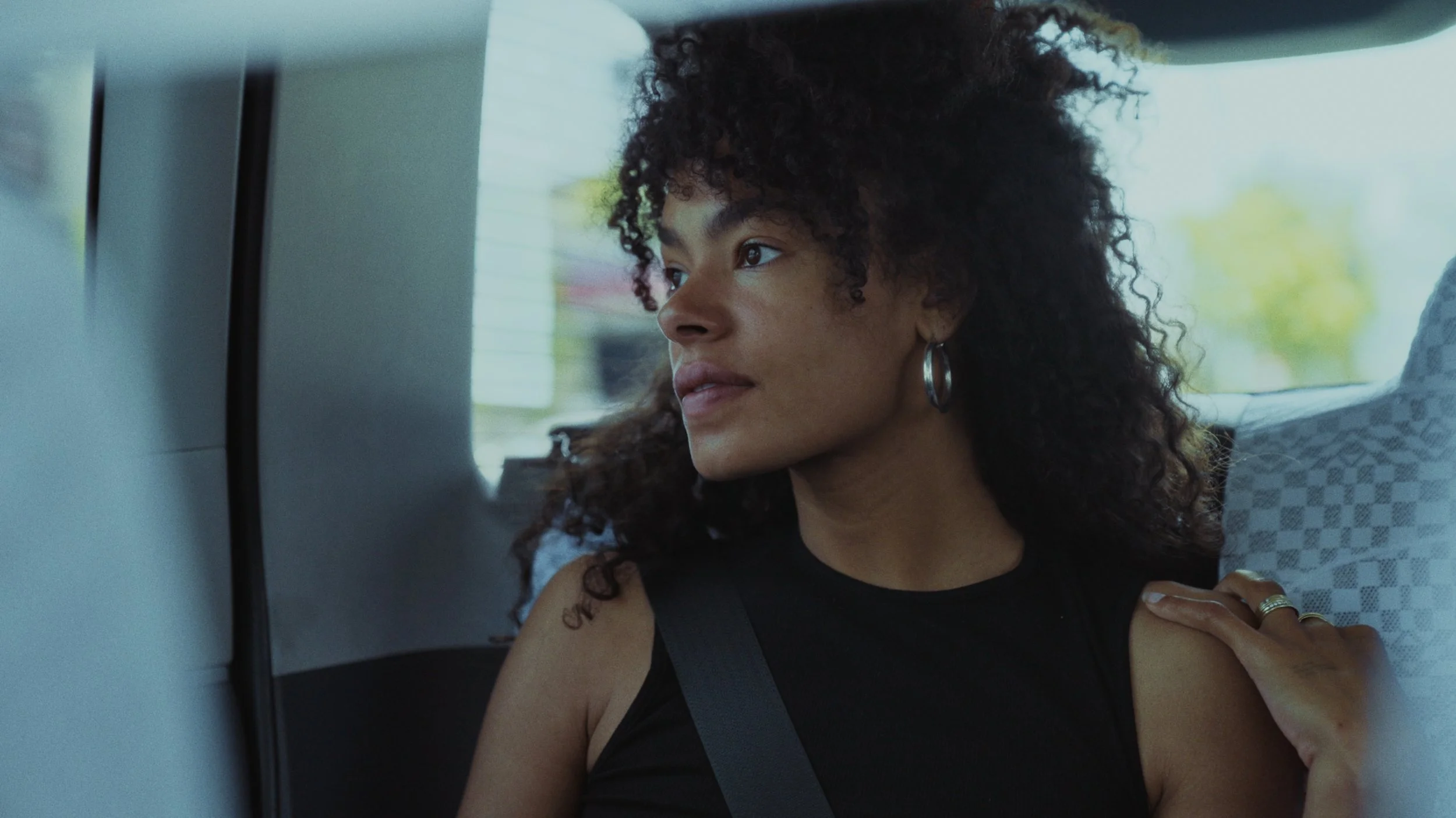 A woman with curly hair wearing a black sleeveless top and hoop earrings, looking out a car window.