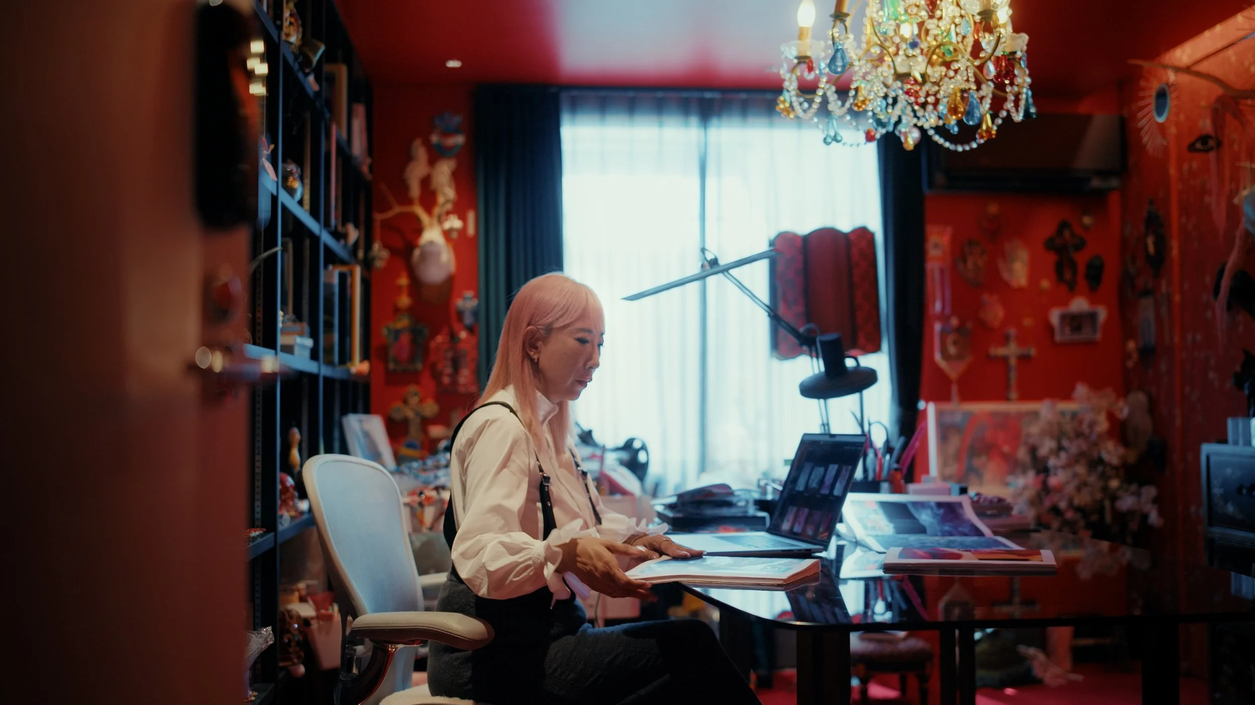 A woman with light pink hair sitting at a desk, reading a book in a room decorated with red walls, floating shelves, and a colorful chandelier.
