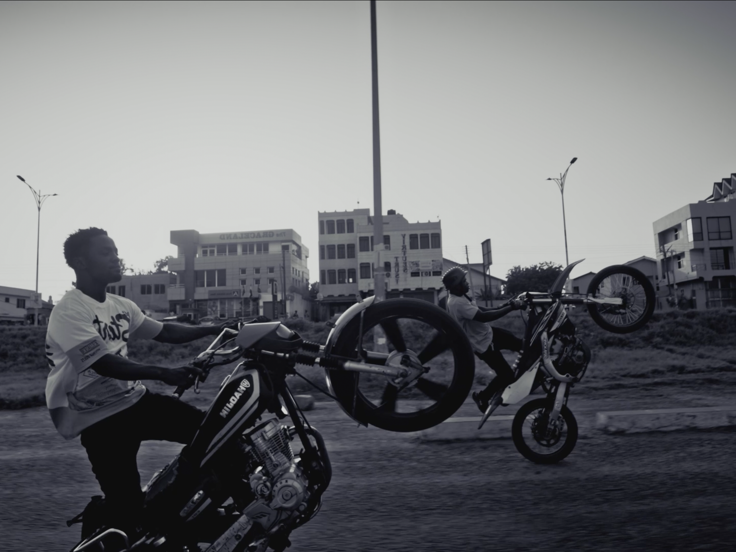 Two men riding motorbikes on a street with buildings and streetlights in the background, black and white photo.