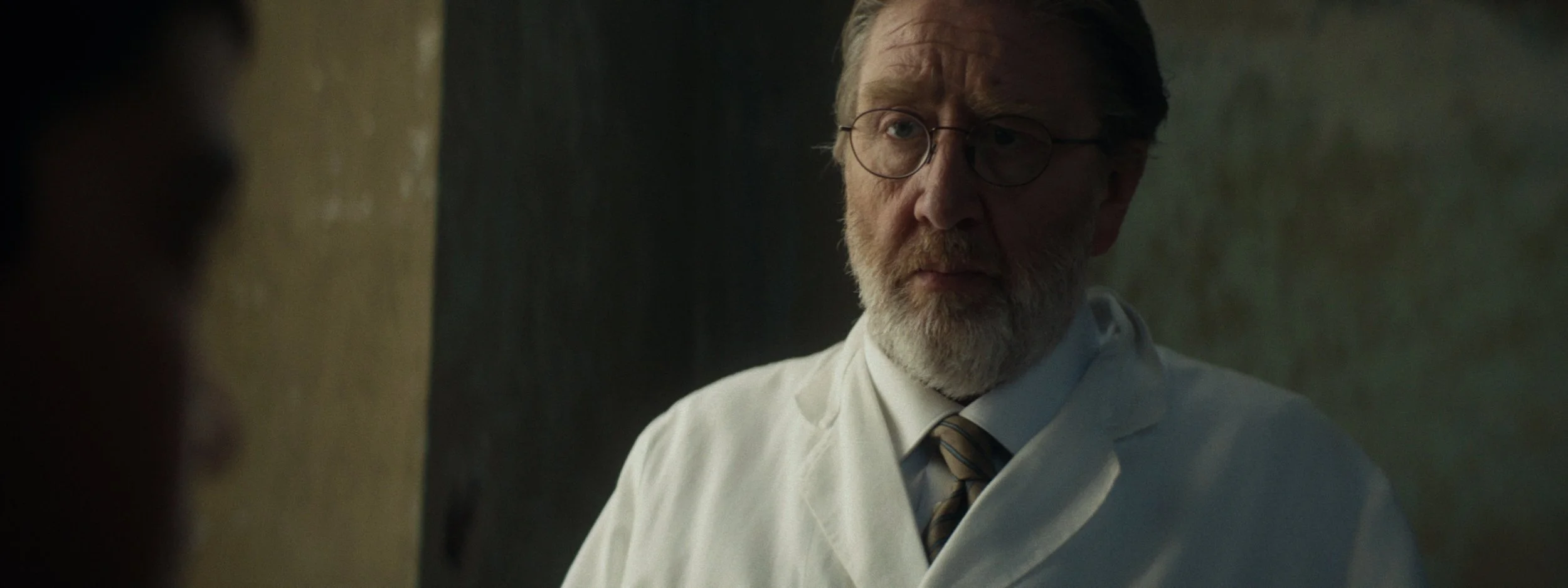 A man with glasses and a beard, wearing a white lab coat and a striped tie, looking serious and pensive.