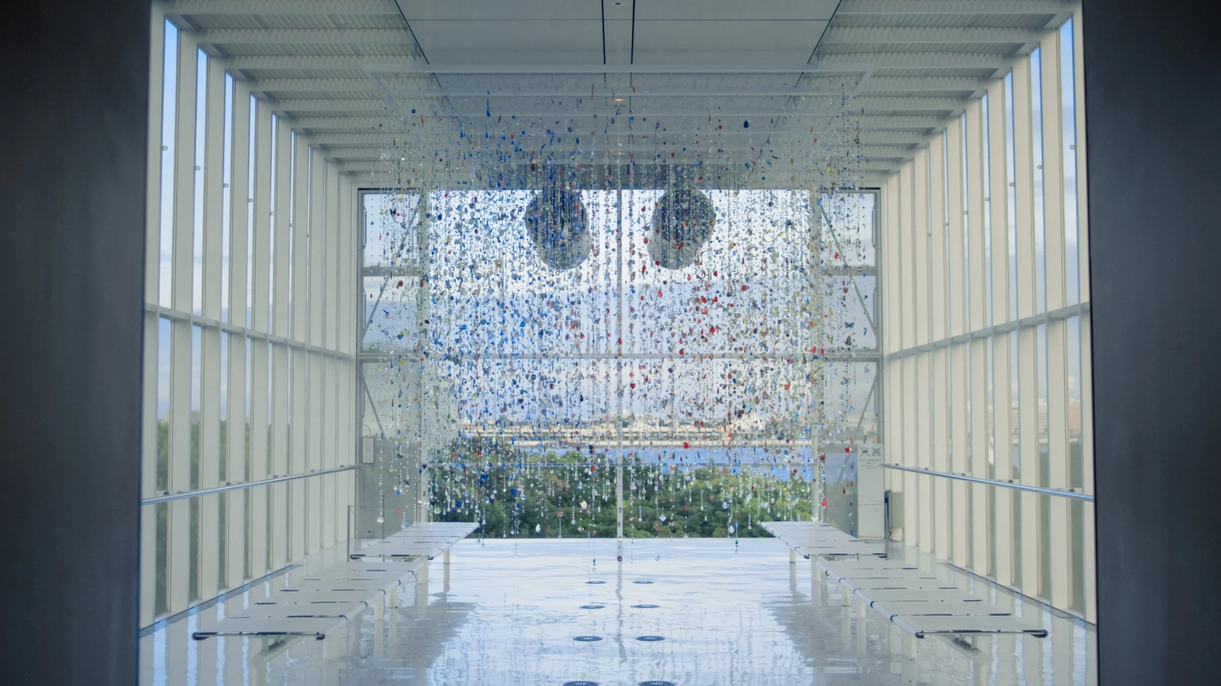 Interior view of a modern building with large windows, decorated with hanging colorful glass art, overlooking an outdoor landscape with trees and a cloudy sky.