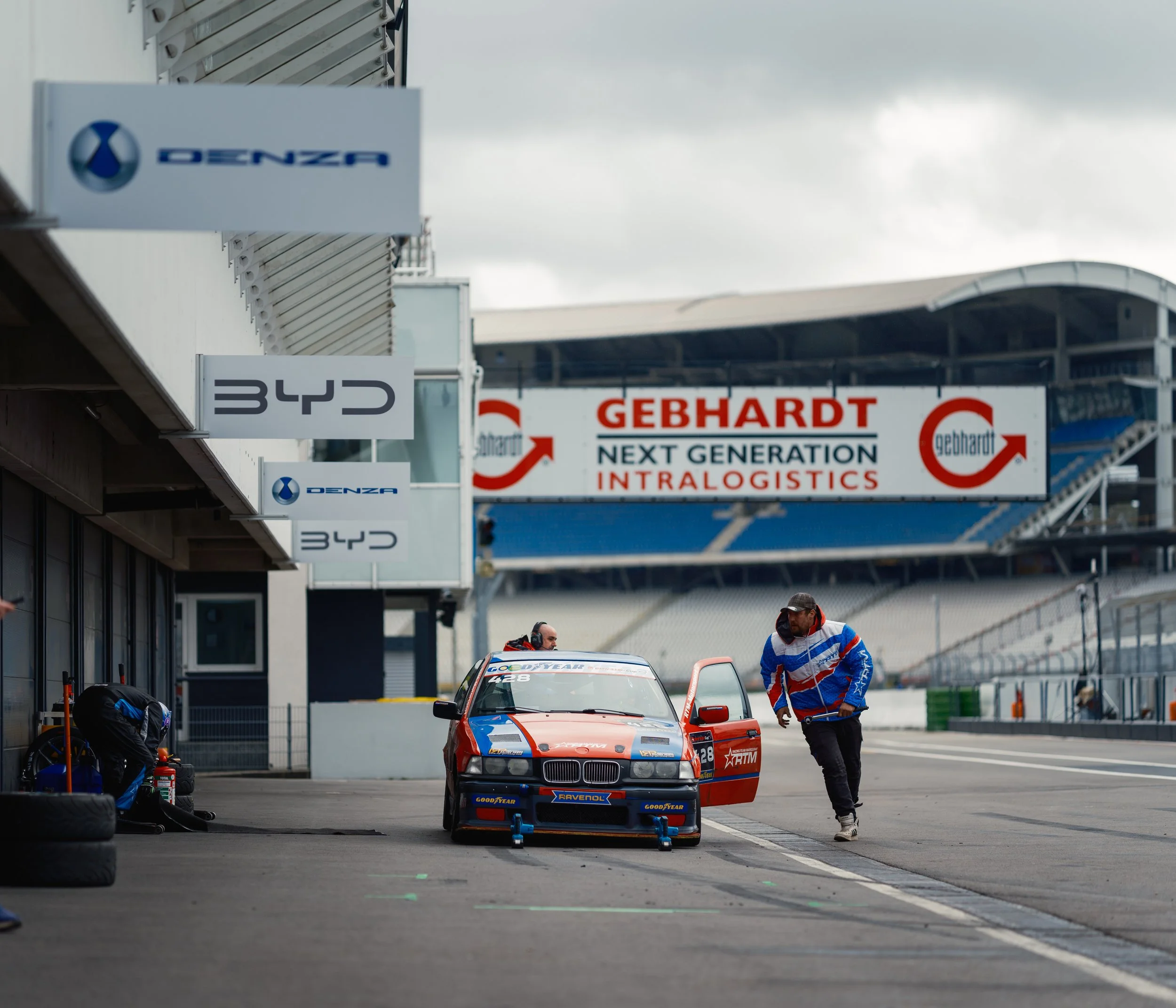 A racecar with a blue, orange, and red color scheme parked in a pit lane at a race track. Two crew members are near the car, one wearing a helmet and the other in a racing suit. The background has pit signs and a large banner with the Gebhardt logo a