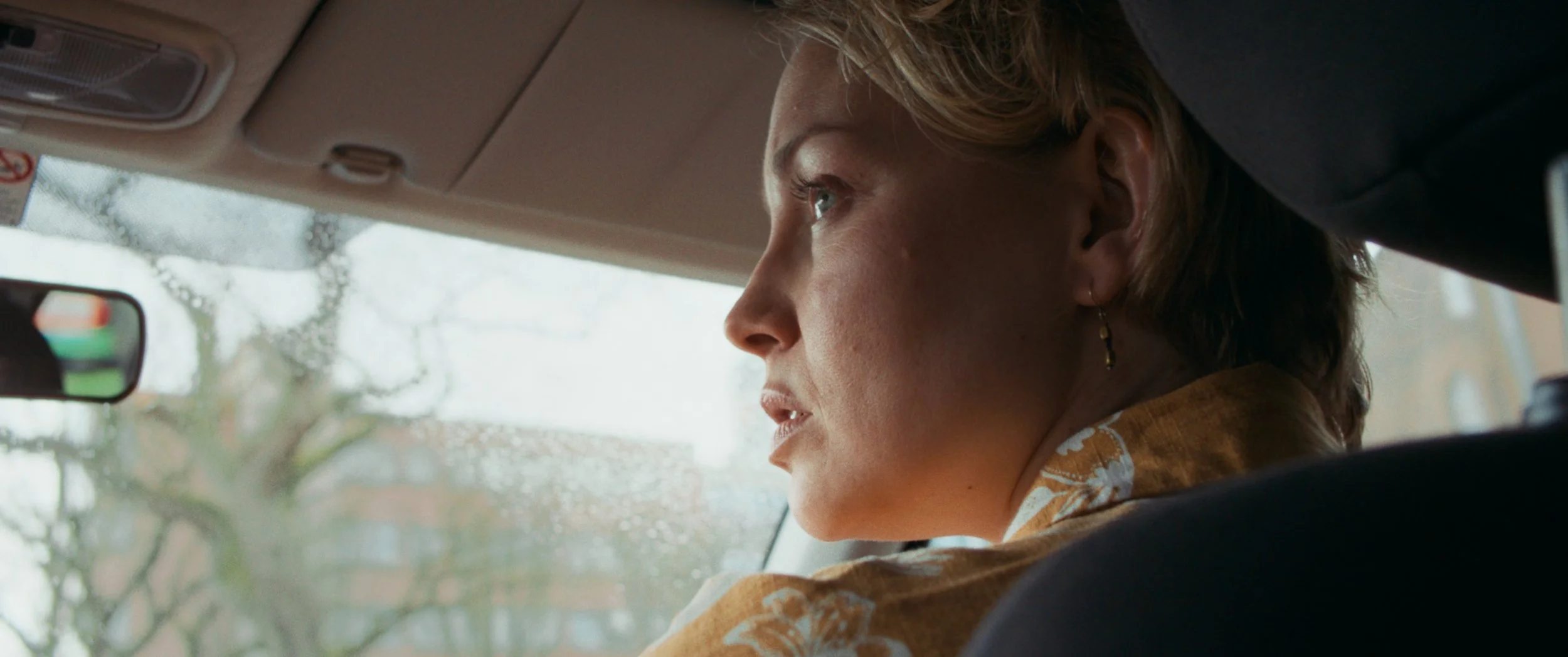 A woman with short hair and earrings sitting in the passenger seat of a car, looking outside through the windshield during rainy weather.