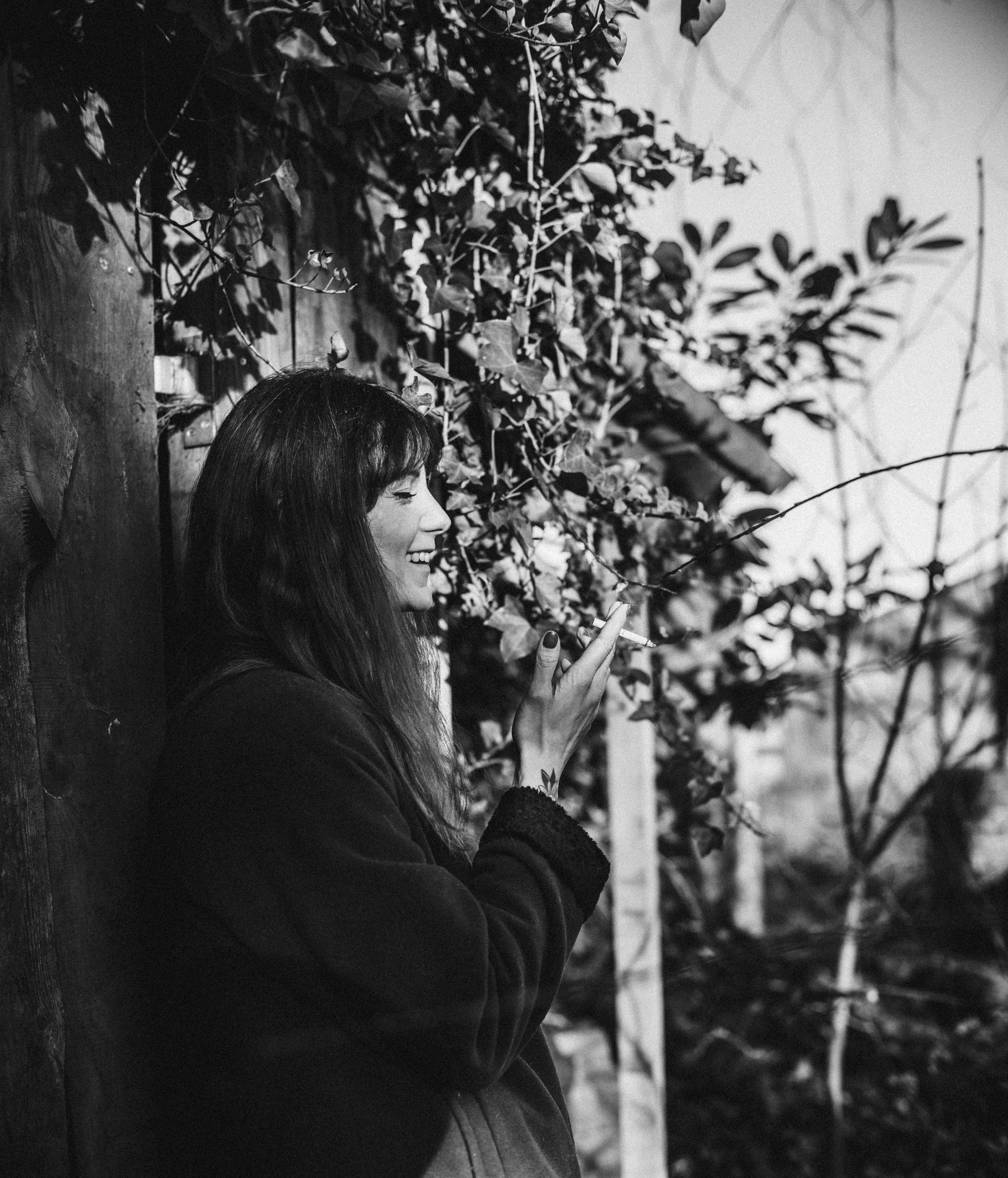 A woman with long hair smiling and looking at her cellphone, standing next to a wooden post covered in climbing plants, outdoors.